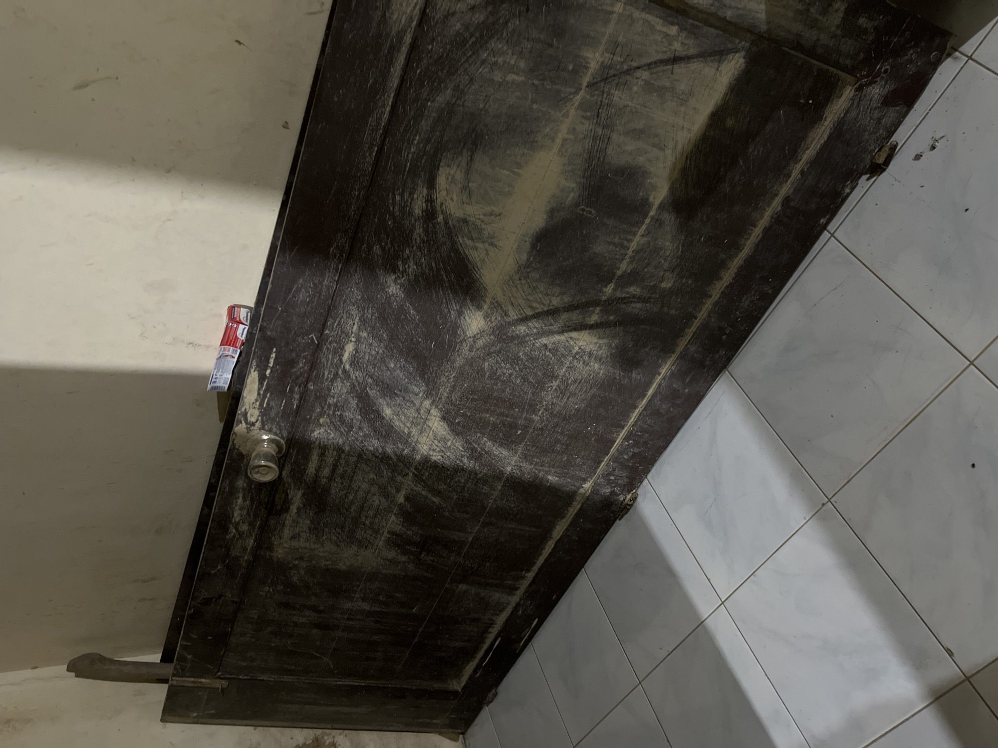 The image shows a closed, dark brown wooden door that appears to be old and weathered. The door is heavily marked with dust and grime, obscuring its original finish. It has a round, clear glass doorknob. A portion of a white tiled wall is visible to the right of the door, with thin gray grout lines. To the left of the door, a beige wall is visible, with some scuff marks and dust. A dark-colored object, possibly a kick plate or a piece of furniture, is resting against the bottom left corner of the door. A small, metallic object, potentially a lock or a hinge, is visible at the bottom right corner of the doorframe. In the upper left section of the image, a small portion of a red and white object, possibly a can or a label, is attached to the door. The lighting suggests an indoor setting, possibly dimly lit, with shadows cast across the door and the tiled wall. The overall impression is one of a neglected or unused space. No people are visible in the image. The scene appears to be static, with no apparent activity taking place. The location context provided is Kota Kuala Simpang, Indonesia.