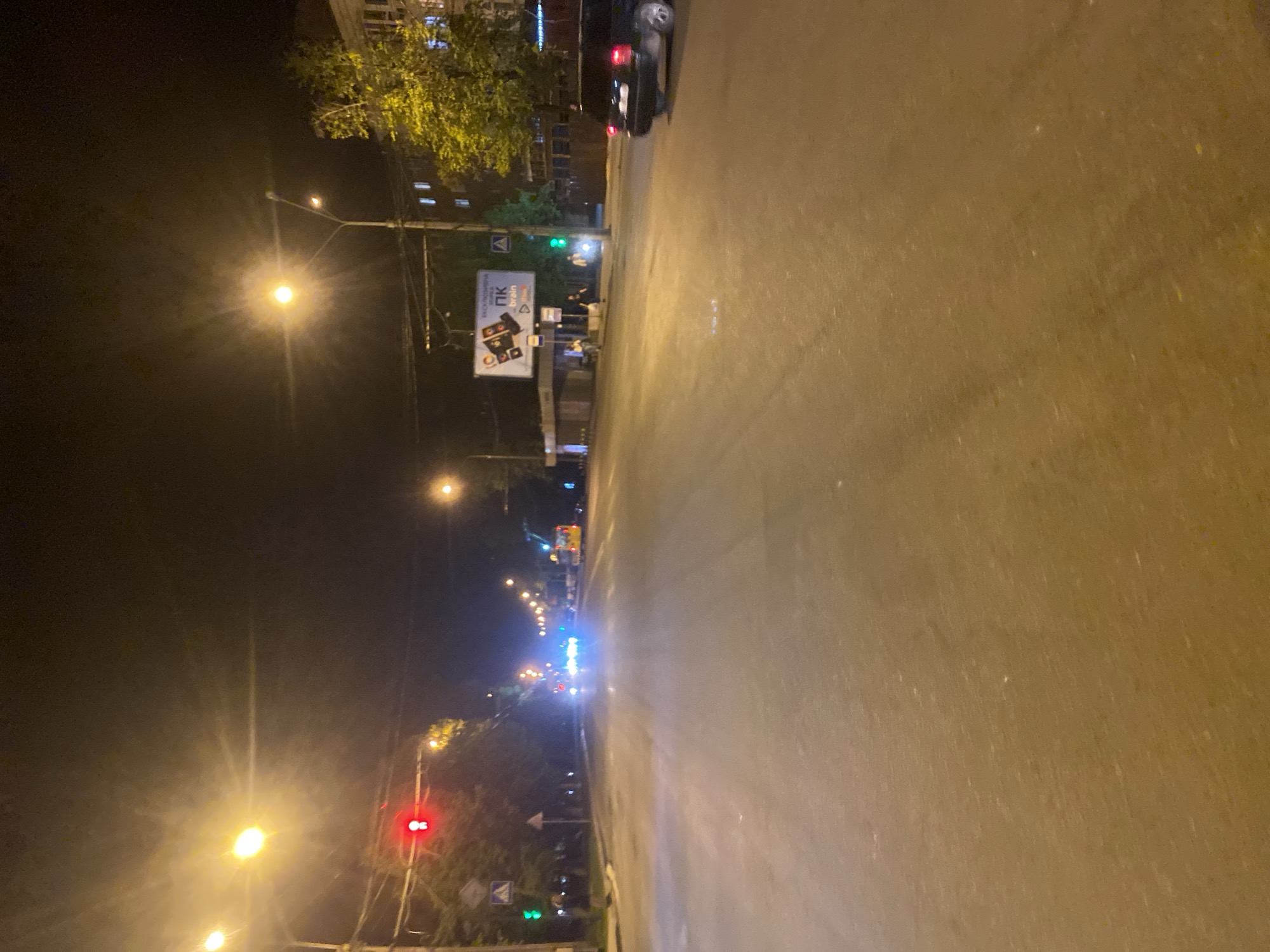 The image depicts a nighttime street scene in Poltava, Ukraine, viewed from ground level looking along a wide, wet asphalt road that stretches into the distance. Streetlights illuminate the scene, casting a golden glow on the road. A black car is parked on the left side of the road at the top of the frame. Further down the street, there are buildings with lit windows, trees, traffic lights (one showing red), and street signs. A large billboard is visible on the left side of the road near the parked car. No people are readily visible in the immediate foreground. The image suggests a quiet, possibly late-night or early-morning setting.