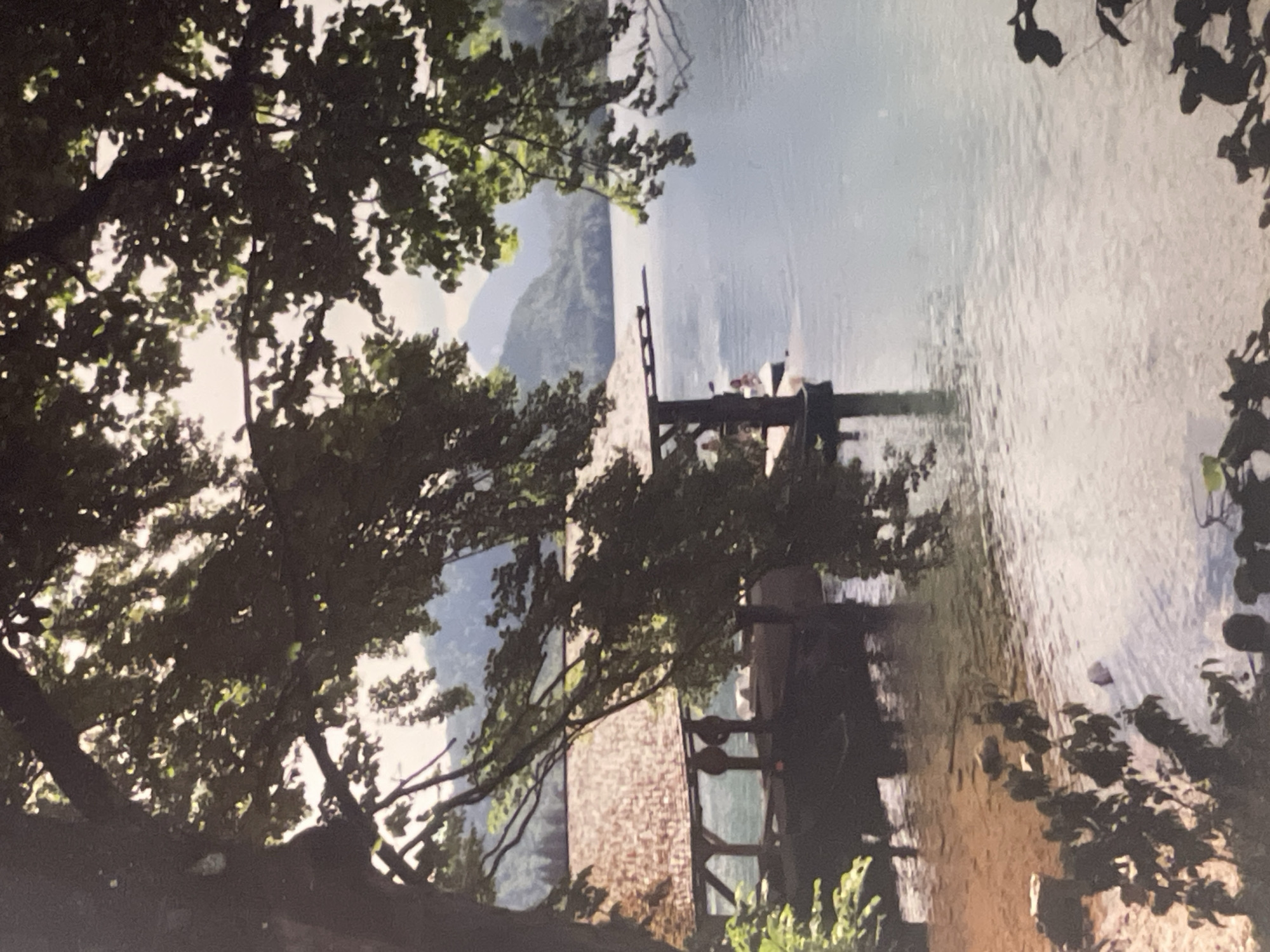 The image depicts a lake scene framed by trees. A small pier extends into the water, with two figures visible on it. The shoreline is sandy. In the background, mountains are discernible. There is no indication that this location is in Maidstone, United Kingdom. Further information is needed.