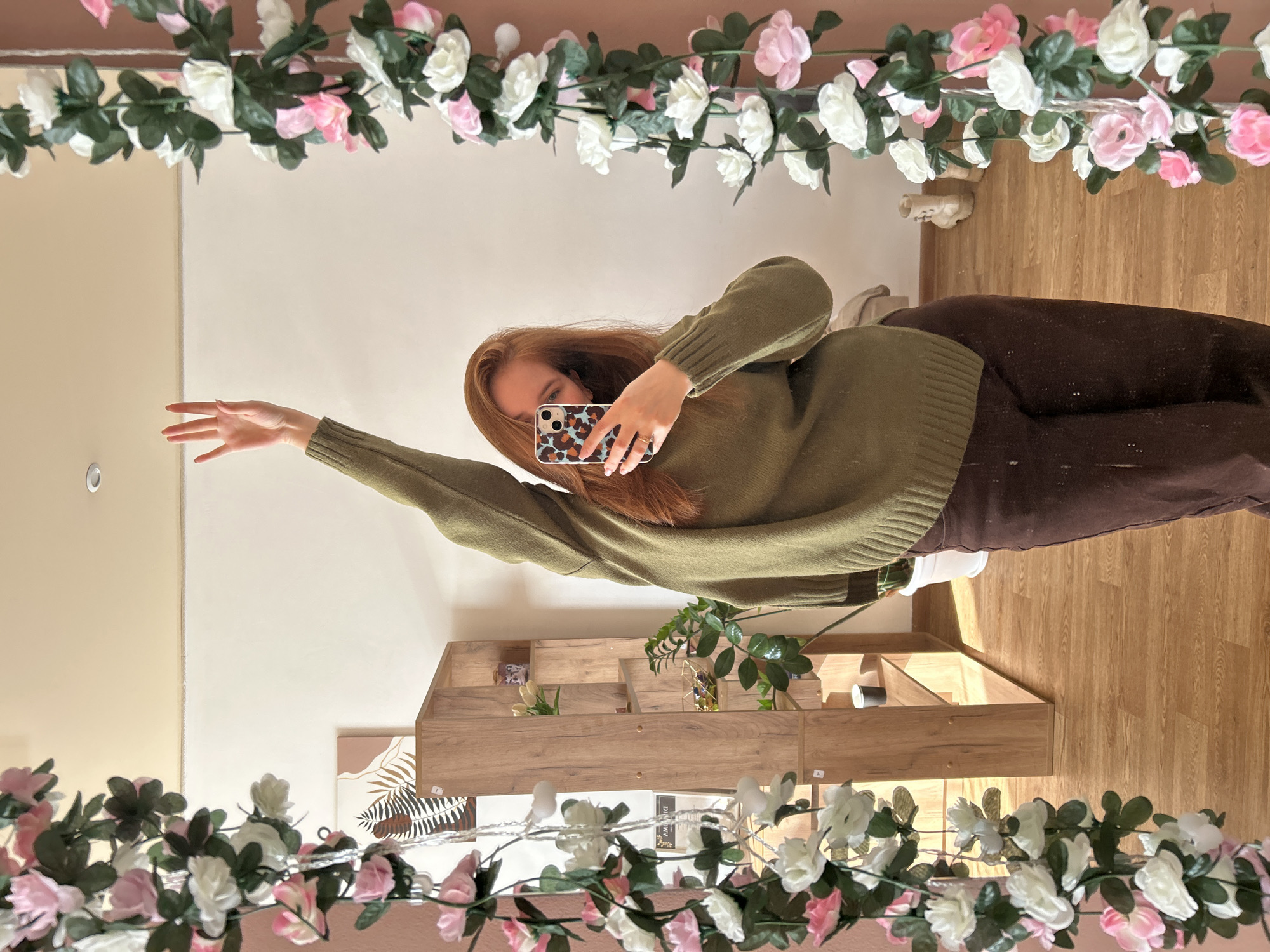 A young woman takes a selfie in front of a mirror decorated with artificial flowers. She wears an olive-green sweater and brown pants. Her right arm is raised. The background includes a shelf with decorations, a wall, and a wooden floor. The image might be taken indoors, possibly in a home. There is no definitive indication that this image was taken in Chaikovsky, Russia.