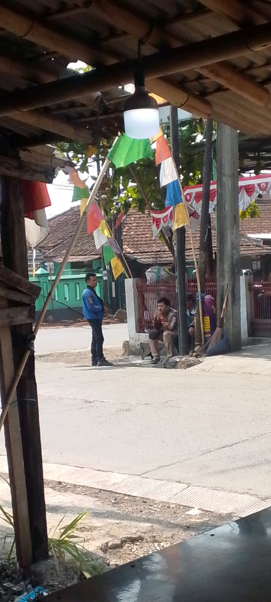 The image depicts a street scene in Cilaku, Indonesia, under a bamboo-like overhead structure. In the foreground, the edge of a dark reflective surface is visible, possibly a table or counter, with some green foliage and debris beneath it. A wooden support pillar is on the left.
The middle ground shows a paved road with a textured concrete curb. On the sidewalk, a man in a blue jacket and black pants stands facing to the right. To his right, two men are seated. One man, wearing a brown and grey striped shirt and dark pants, is holding something to his mouth, possibly food or a cigarette. Another person, partially obscured, sits to his right. Behind them, a purple bag and a broom are leaning against a pole.
In the background, traditional Indonesian houses with tiled roofs are visible. A bright green building with white-trimmed windows is on the left. Colorful flags, including red and white, are strung across poles and trees, suggesting a festive occasion or national celebration. A pole with multiple small, colorful pennant flags is prominently displayed in the center. The overall atmosphere appears to be casual and outdoors.