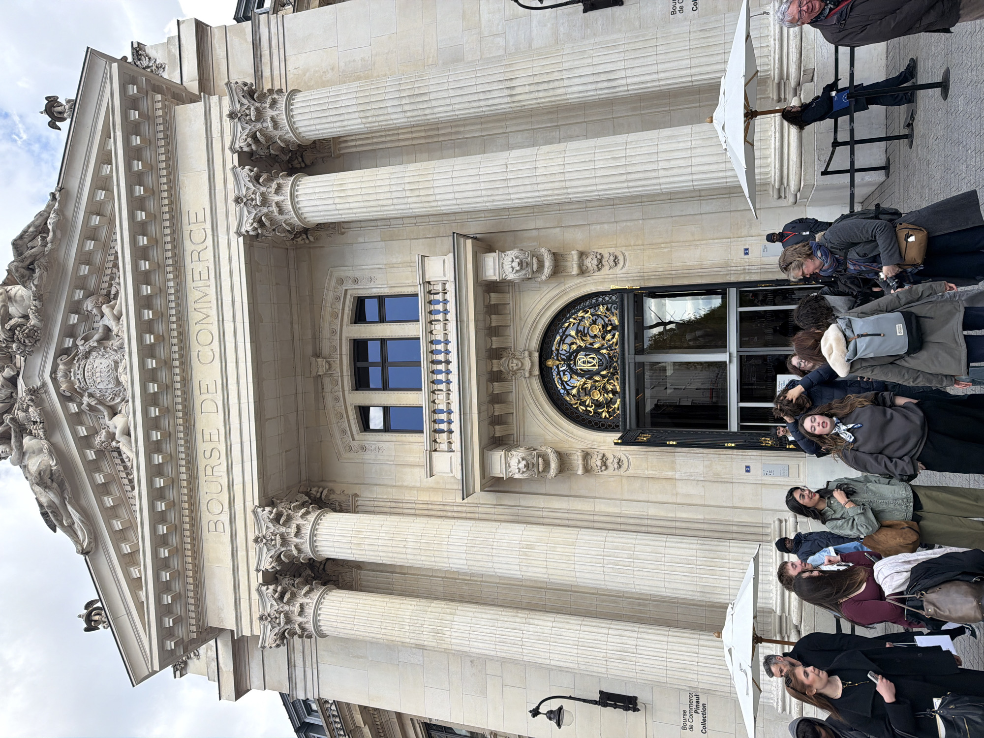 The image depicts the front facade of the Bourse de Commerce – Pinault Collection building in Paris, France. The grand, neoclassical stone structure features a prominent entrance framed by six fluted Corinthian columns supporting an ornate pediment adorned with classical sculptures, including reclining figures and cherubs. The words "BOURSE DE COMMERCE" are engraved above the central columns.
Below the pediment, a central arched entryway is visible, featuring dark, ornate wrought-iron gates with gold accents, one of which displays an intertwined "PC" monogram. Above this entrance is a small balcony with a stone balustrade and a window with dark frames. The facade is further embellished with stone carvings, including lion heads above the archway. Wrought-iron lamps are mounted on the wall to either side of the entrance. A sign on the left column identifies the building as "Bourse de Commerce Pinault Collection."
In the foreground, a diverse group of approximately 20-30 people are gathered on a paved stone surface directly in front of the building. Most individuals are standing in small clusters, conversing or observing their surroundings. Two large white commercial umbrellas with wooden poles are positioned on either side of the entrance, providing shade. On the right, black stanchions with connecting ropes define a queuing or restricted area. Individuals wear varied casual to smart-casual attire. Notable individuals include a woman in a black coat holding a smartphone, a woman in a green jacket with glasses and a brown bag, and an older man in a brown jacket on the far right. The sky above is overcast with visible cloud formations.