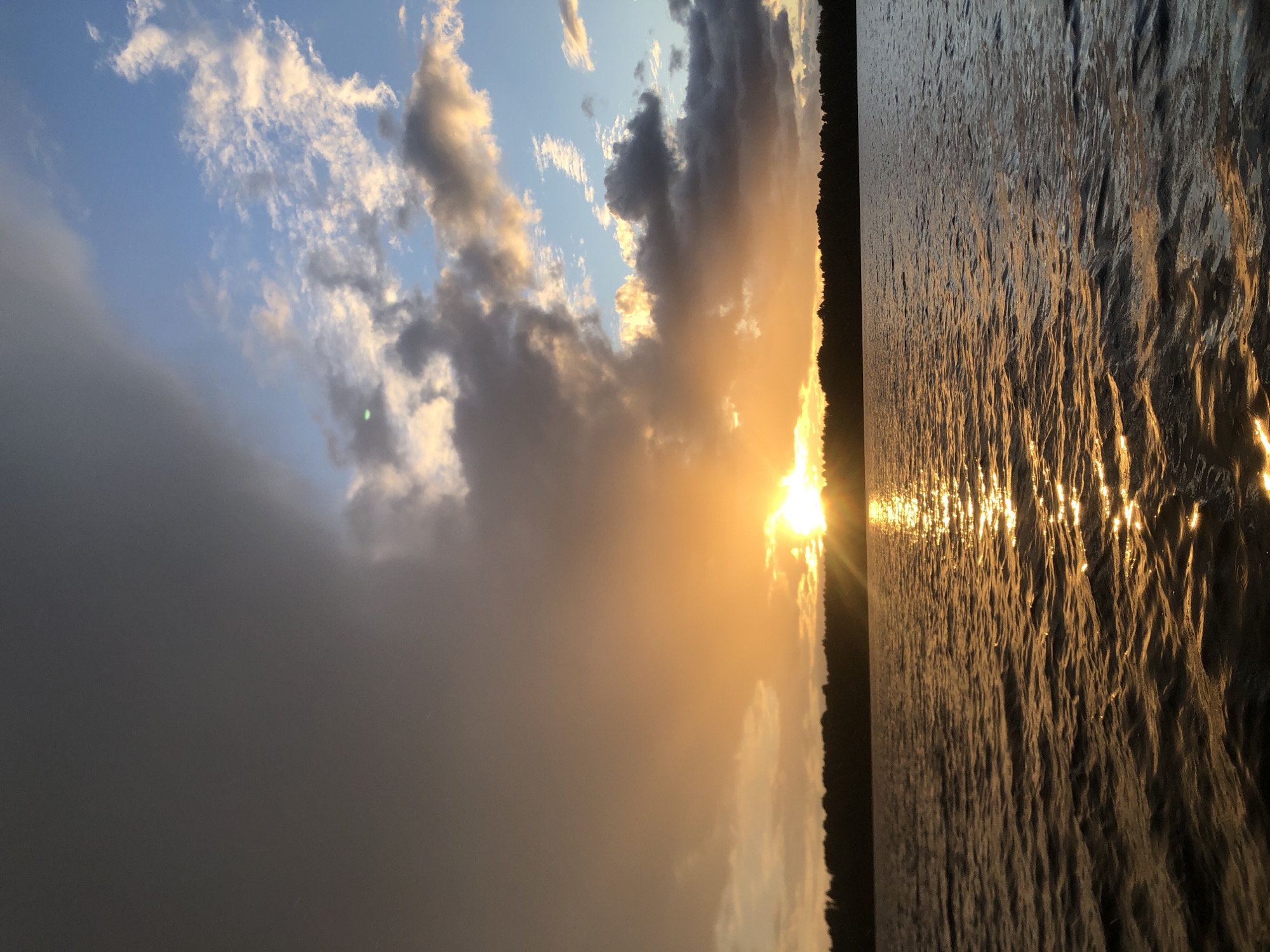 A horizontal photograph captures a sunset over a body of water, likely a lake or river, viewed from a low angle. The sun, a bright orb, is partially obscured by dramatic, dark grey and wispy white clouds tinged with golden light. The sky transitions from a deep blue at the top left to a soft orange and yellow hue near the horizon.
To the right of the clouds, the sun's rays burst forth, casting a shimmering golden reflection across the rippled surface of the water. The water occupies the right two-thirds of the frame, its texture characterized by small, rhythmic waves. Along the left horizon, a dark silhouette of trees forms the edge of a landmass.
The scene suggests a serene and possibly tranquil moment, illuminated by the warm glow of the setting sun. The overall impression is one of natural beauty and the peaceful transition from day to night. The image appears to be taken outdoors, possibly from a boat or a shoreline, in Svirstroy, Russia. The time of day is clearly evening, as indicated by the sunset. There is no visible text in the image.