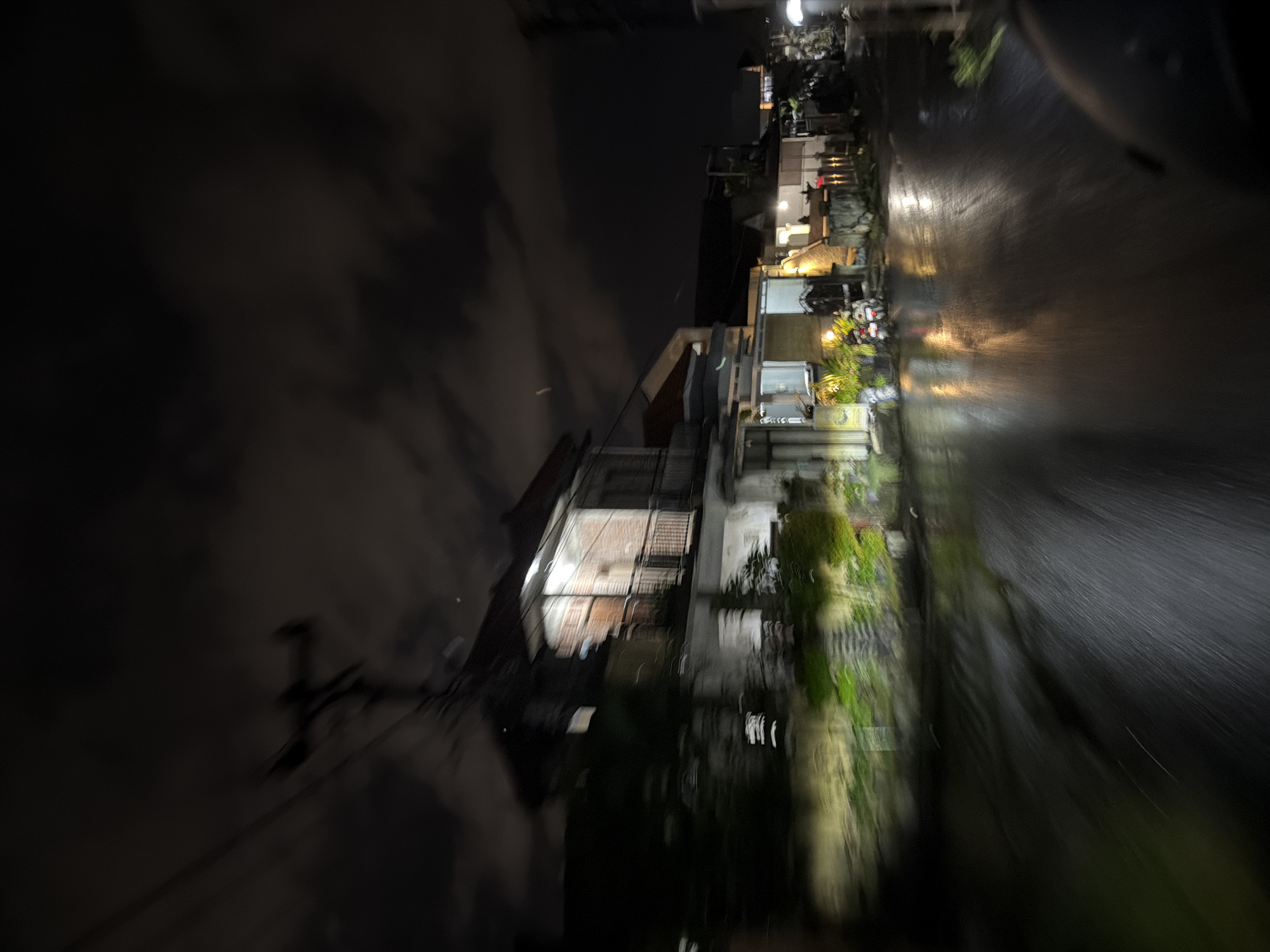 This blurred image captures a nighttime street scene, likely taken from a moving perspective. The setting appears to be a residential or commercial area, with various buildings lining one side of a wet street.
The time of day is dark night, with the sky displaying faint cloud patterns on the left. Artificial light sources, including bright white and warm yellow lights, emanate from within and around the buildings, illuminating parts of the scene and creating reflections on the street. These structures vary in size, some appearing to be multi-story with visible windows and balconies. Greenery, such as plants and trees, is interspersed along the roadside and in front of the buildings, exhibiting motion blur.
The street surface is notably wet and reflective, suggesting recent rain or a damp environment. Streaks of light from headlamps or other sources are visible on the wet pavement, contributing to the strong motion blur evident across the lower and left portions of the image. A motor vehicle, possibly a scooter, is partially discernible parked near one of the buildings, indicated by a faint red light reflection. Utility poles and overhead wires are faintly silhouetted against the dark sky on the far left.
The overall image is characterized by significant motion blur, indicating a fast shutter speed combined with movement during capture, or a longer exposure from a moving platform. The scene evokes a quiet, nocturnal atmosphere. No discernible text is visible. The location context indicates this scene is in North Kuta, Indonesia.