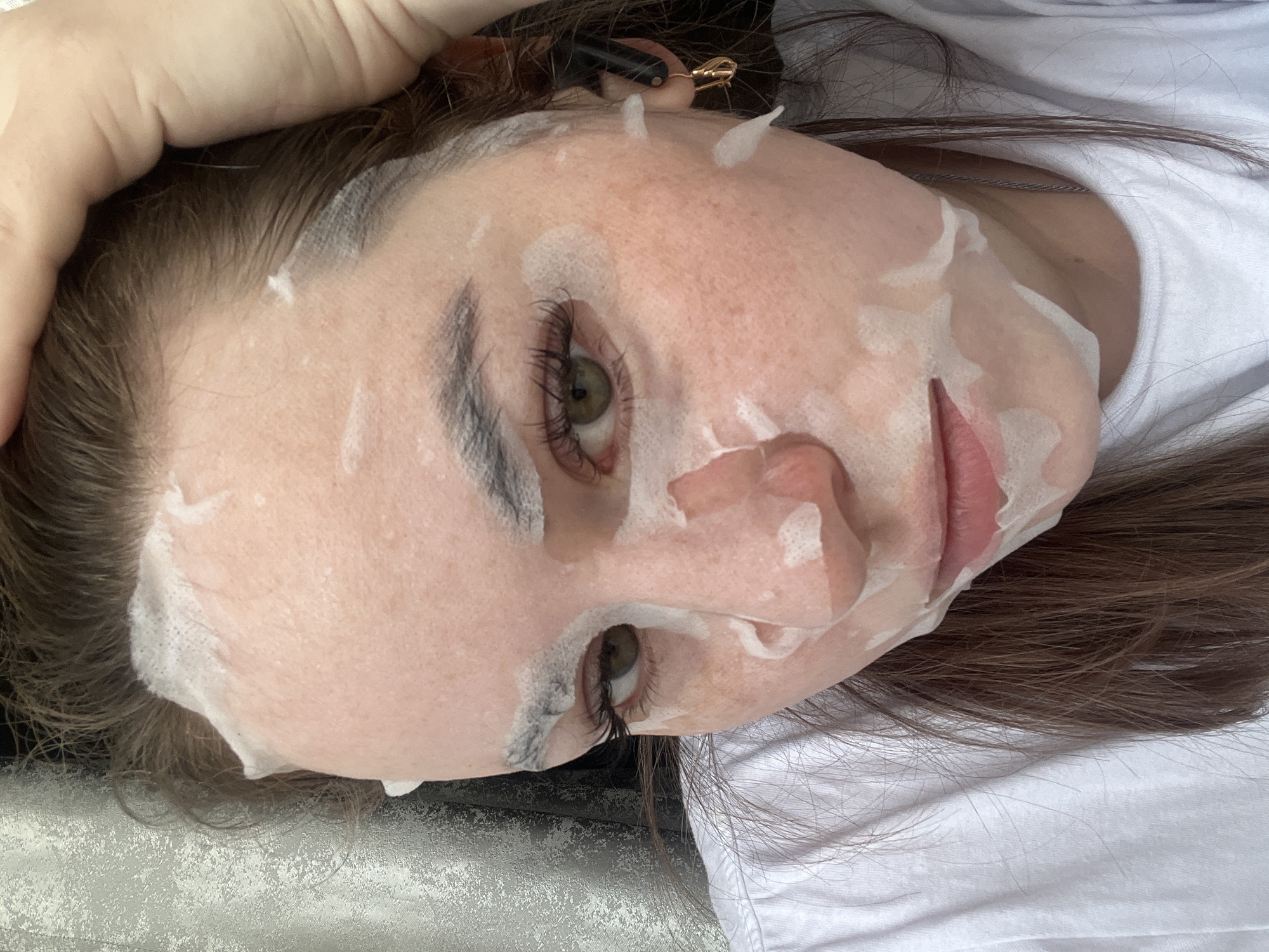 The image, captured in Chaikovsky, Russia, presents a close-up, angled view of a person's face undergoing a beauty treatment. The subject, appearing to be female, is lying down indoors, with her head slightly tilted. Her face is covered with a white sheet mask or a similar facial treatment, with openings for her eyes and mouth. White, foamy residue from the mask is visible along the edges, particularly on her forehead and temples, blending into her hairline.
Her eyes are open, revealing light-colored irises, possibly greenish-hazel, and long, dark eyelashes. Her eyebrows are dark and well-defined. Her lips, visible through the mask's opening, are slightly parted and appear to have a natural pinkish tone. Her complexion is fair.
Her brown hair frames her face and extends onto a white surface, which could be a pillow, bed, or a treatment table. She is wearing a white t-shirt or similar garment, parts of which are visible. A light-skinned hand is positioned near the top left of her head, and a small, gold and black hair clip can be seen in her hair near her temple. A thin, metallic necklace is also visible around her neck. The overall impression is one of relaxation, common during a personal care or spa treatment. There is no visible text in the image.