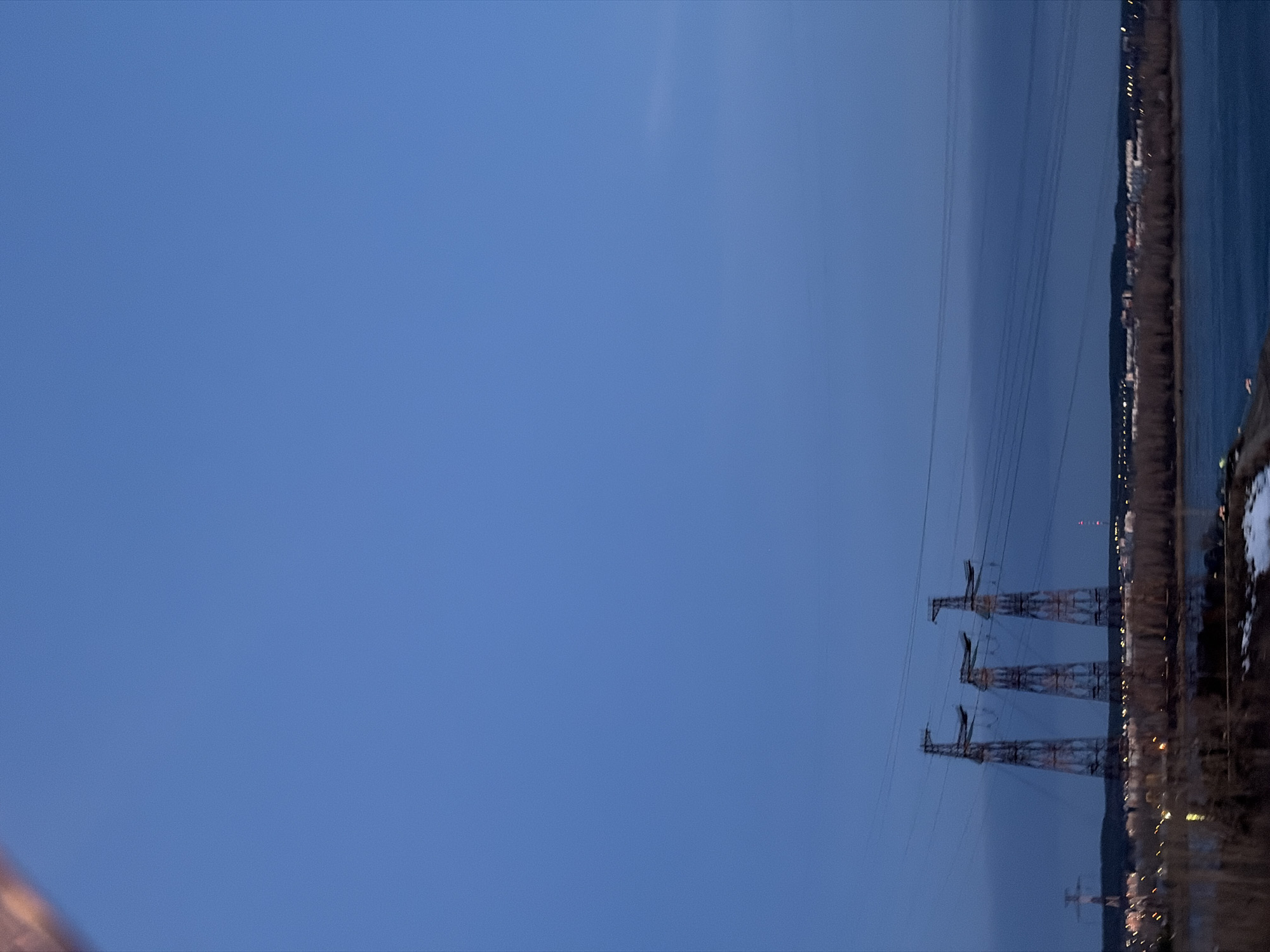 The image depicts a night scene in Chaikovsky, Russia. Three electrical pylons stand in a row, carrying power lines. A body of water is located to the right of the pylons, with a shoreline visible beyond. Lights suggest a settlement or city running along the shore. The sky is a dark, dusky blue.