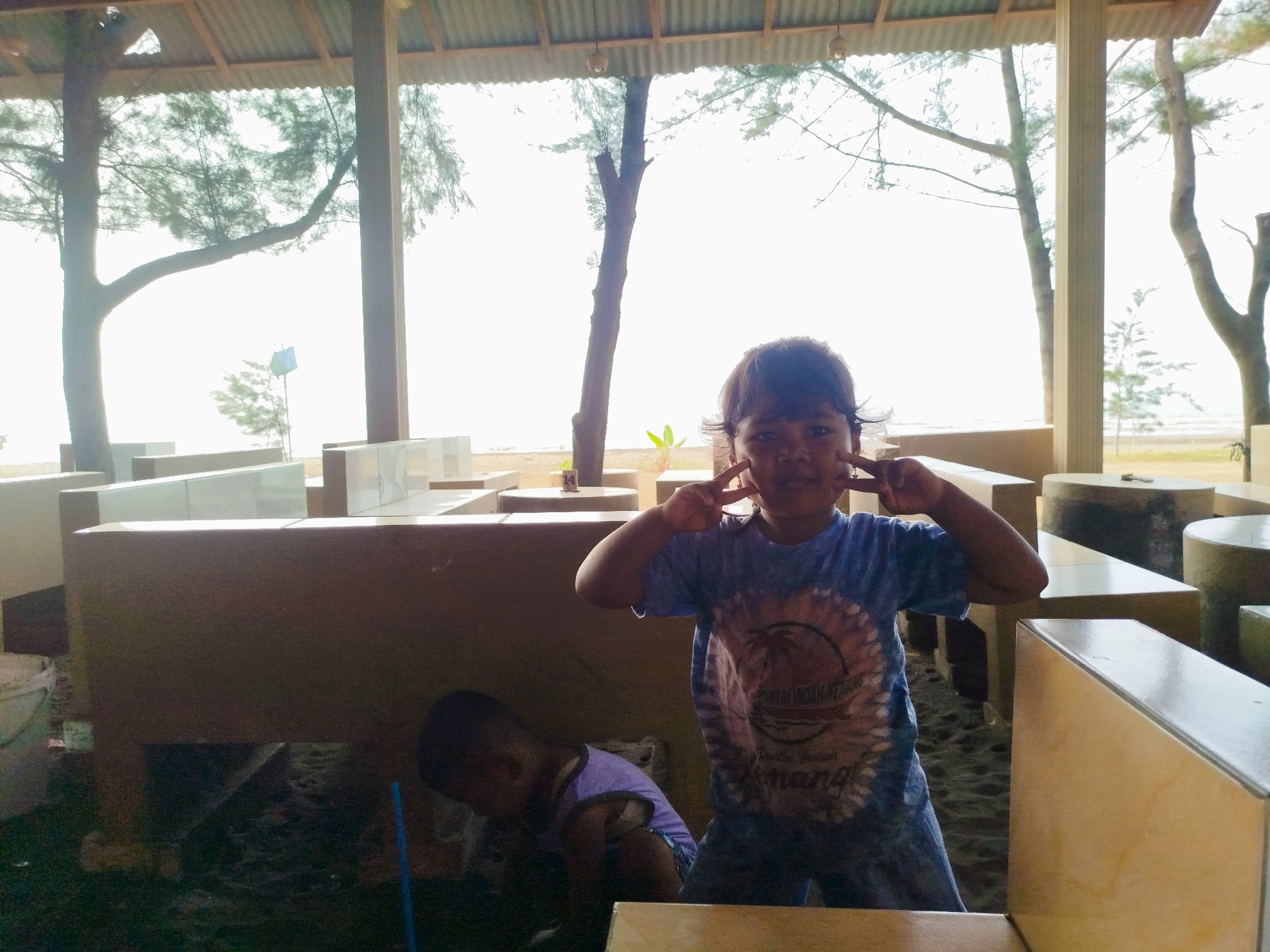 In Jungsemi, Indonesia, a girl wearing a blue t-shirt with a circular graphic of a palm tree and waves stands on sandy ground, striking a peace sign pose with both hands near her face. A young boy in a purple tank top is crouched to her left. They are situated outdoors under a corrugated metal roof supported by wooden posts. In the background, trees and a bright, overcast sky are visible, suggesting a beachside location. The scene appears to be a casual outdoor seating area.