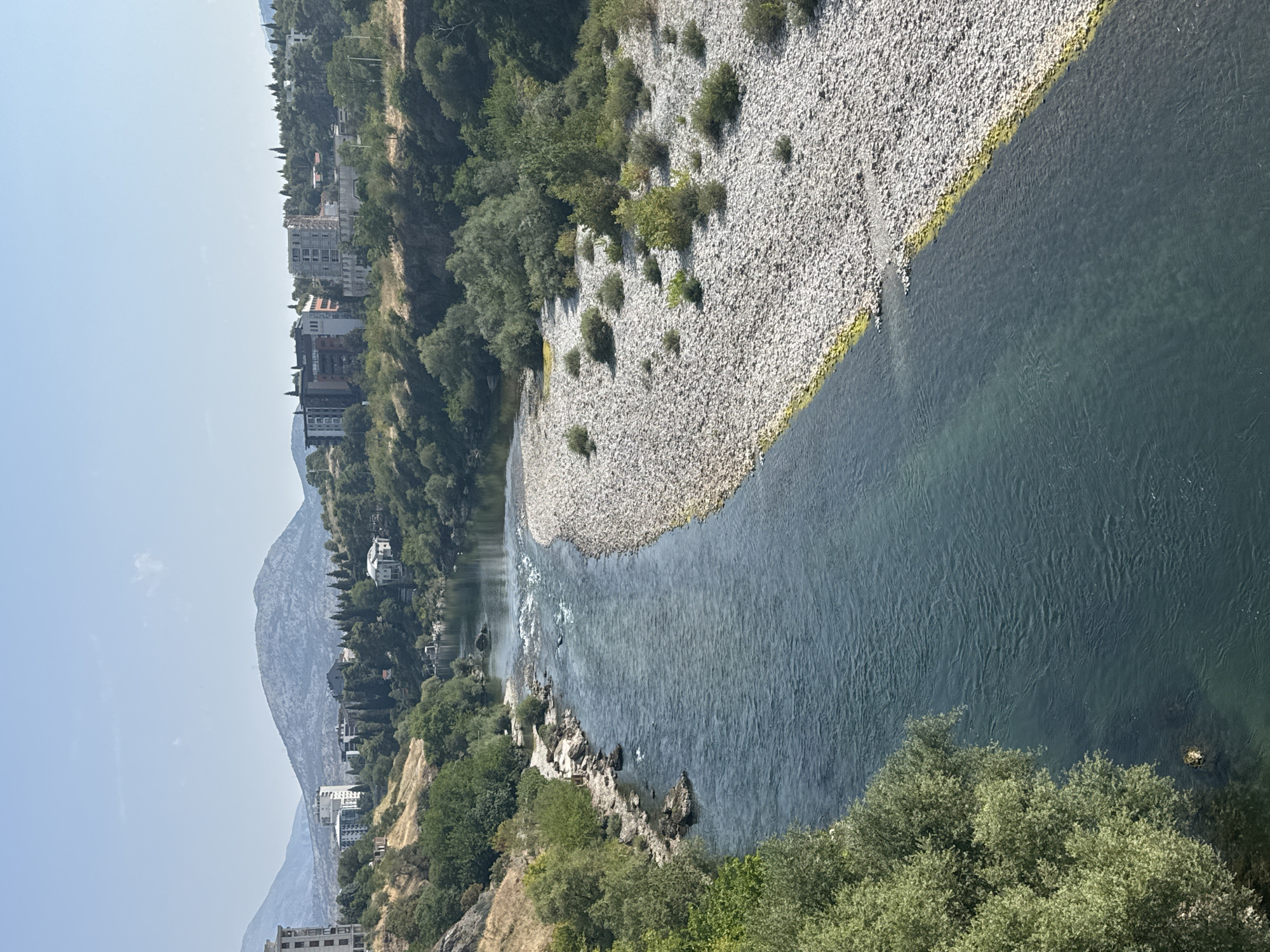 This is an aerial shot of the Moraca River in Podgorica, Montenegro, taken on a clear day with the sun high in the sky. The river flows from the background towards the foreground, with a wide, shallow section of rocks and pebbles on the right bank and a deeper, darker blue channel on the left. Lush green trees and vegetation line both banks of the river. In the background, on the hills overlooking the river, are several apartment buildings and residential structures, interspersed with more trees. Further in the distance, rolling hills and mountains form the backdrop under a pale blue sky. The scene is peaceful and natural, with a touch of urban development visible on the hillsides.