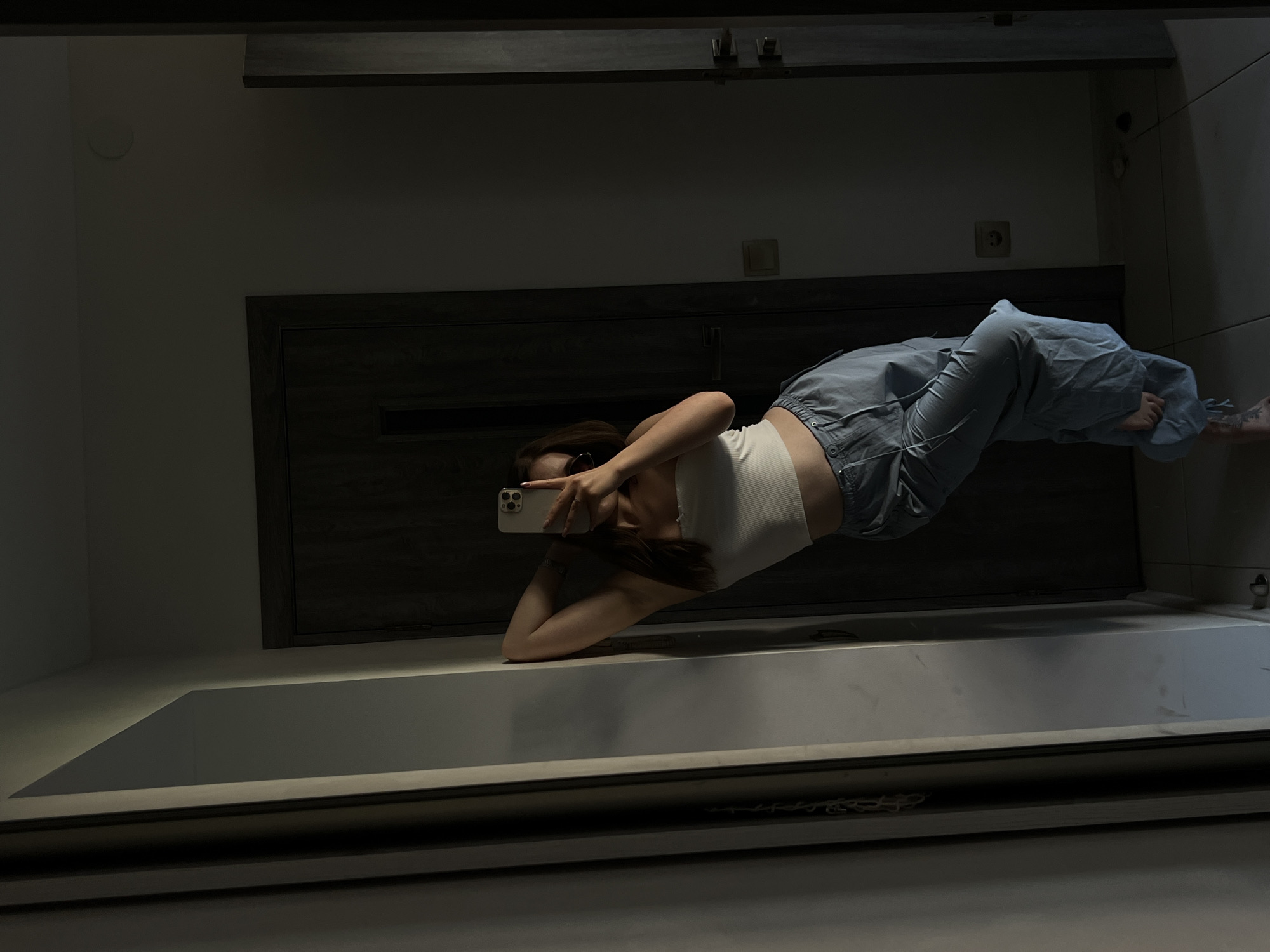 A young woman, possibly in Ternopil, Ukraine, takes a selfie in a hallway with a mirror. She is leaning against a wall, her left arm bent behind her head, and holding a smartphone with her right hand. She is wearing a white bandeau top and light blue cargo pants. The hallway appears to be indoors, with a dark wooden door visible behind her. The lighting is dim, with shadows casting across the scene. The floor is tiled.