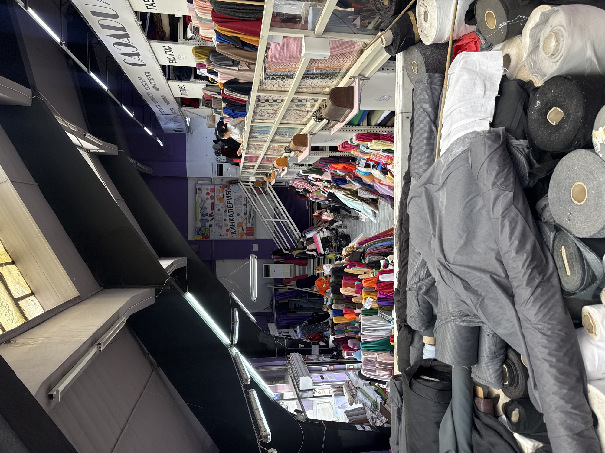 The image depicts the interior of a large fabric store located in Sofia, Bulgaria. The store is a multi-level establishment, with various types of fabrics displayed prominently on shelves and in rolls. In the foreground, large rolls of fabric are stacked, predominantly in shades of grey and black. As the view moves deeper into the store, a wide assortment of colorful fabrics is visible, arranged in neat rows on elevated displays.
On the upper levels, more fabric selections are organized on shelves, featuring a spectrum of colors and patterns. Several individuals are present in the store, interacting with the merchandise and possibly with each other. One group appears to be browsing or discussing fabric choices, while others are engaged in activities behind the counter areas. The store's architecture includes high ceilings with industrial lighting, and staircases provide access to different sections. Signs in what appears to be Cyrillic script are visible on the walls and above the fabric displays, indicating product categories and potentially store branding. The overall atmosphere suggests a bustling retail environment dedicated to the sale of textiles.