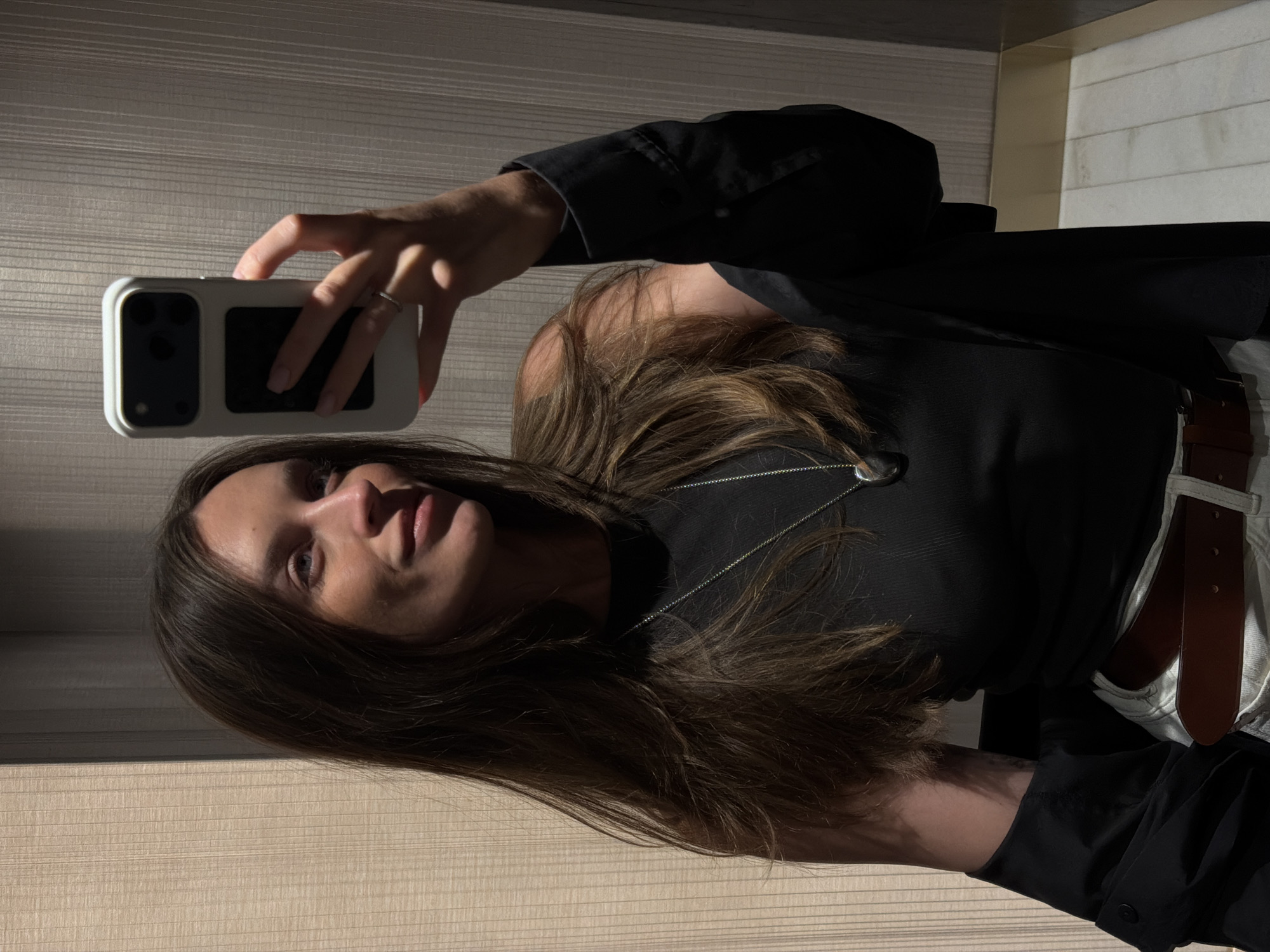 A woman takes a selfie indoors in Budva, Montenegro, using a smartphone. She has light brown hair, wears a black top, and a dark blazer, with a silver necklace visible. A brown belt is also visible. The background shows horizontally aligned panels.