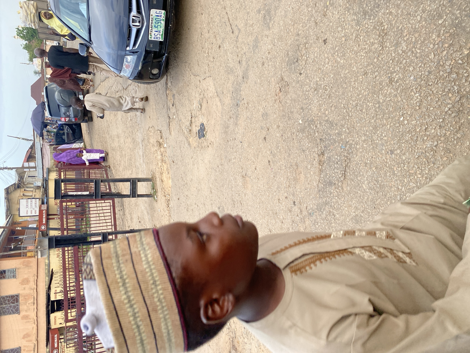 The image shows a street scene in Jos, Nigeria. In the foreground, a young boy wearing a traditional tan kaftan and a striped cap is looking to his right. In the background, several people are walking on the dirt road. A black SUV and a black sedan are parked on the side of the road. The buildings in the background appear to be shops and residential homes. The sky is overcast.