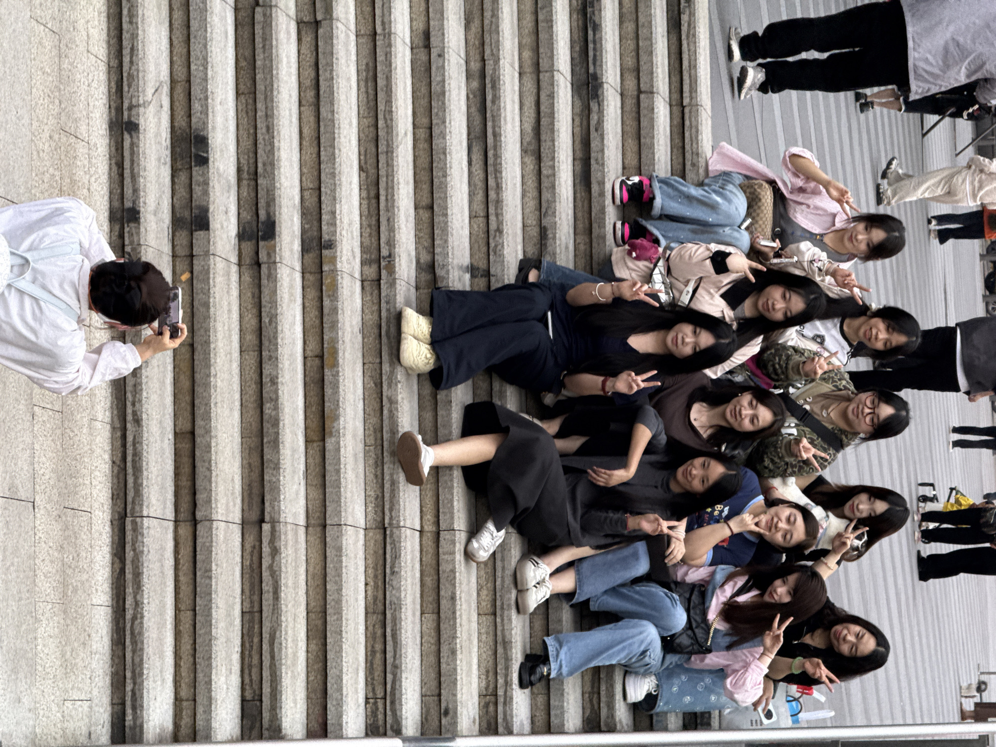 A man in a white shirt takes a photograph of approximately eleven women lying on a stepped surface. The women are stacked together, smiling, and flashing peace signs. The setting appears to be an outdoor public space with a concrete staircase or tiered platform. Yuzhong District, China, cannot be confirmed based on the image.