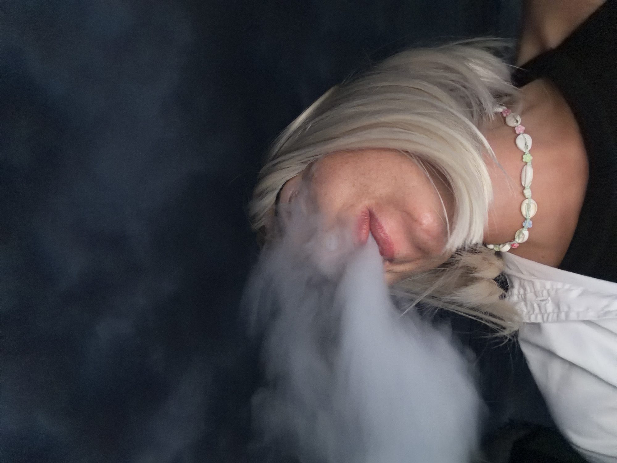 The image shows a close-up, low-angle, outdoor shot of a person with blonde hair exhaling a cloud of white vapor. The person's face is partially obscured by the vapor, with only their mouth, nose, and part of their cheek visible. They are wearing a necklace made of shells and small colorful beads. They are also wearing a white collared shirt partially open over a dark-colored garment. The background is dark and appears to be a night sky with some indistinct cloud formations. There is no visible indication of the specific city of Sochi, Russia, in this image.