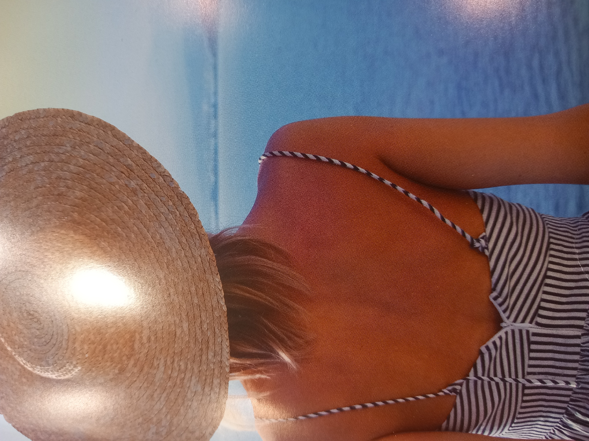 A close-up, sideways shot shows the back of a person wearing a straw hat and a striped bathing suit. The hat is large and woven, with a bright reflection on its surface. The person's skin is tanned, and their hair is light brown and catches the light. The background is a blurred, bright blue, suggesting an outdoor setting like a beach or by the water. The lighting appears to be bright, possibly indicating daytime. There is no visible text in the image. The scene evokes a feeling of relaxation and summer.