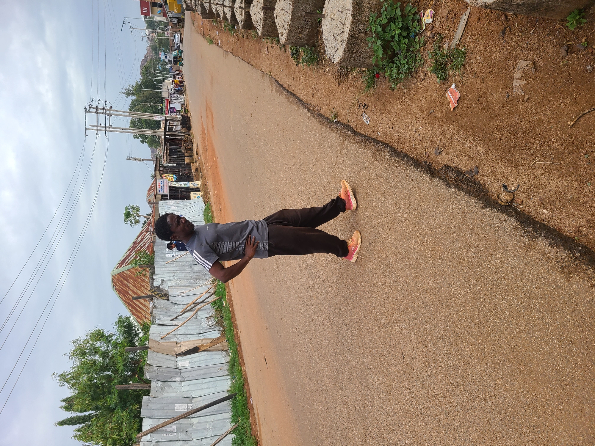 A man in a gray shirt with white stripes on the sleeves and dark pants stands on a paved road. He is facing towards the left side of the frame, with his body nearly horizontal in the image. He wears orange sneakers. The man is on a road in Jos, Nigeria.
On the left, there is a fenced-off area with a corrugated metal roof and vegetation. In the distance, utility poles and other buildings are visible. The sky is overcast. The road is lined with concrete structures. The road has dirt and debris along the edge.