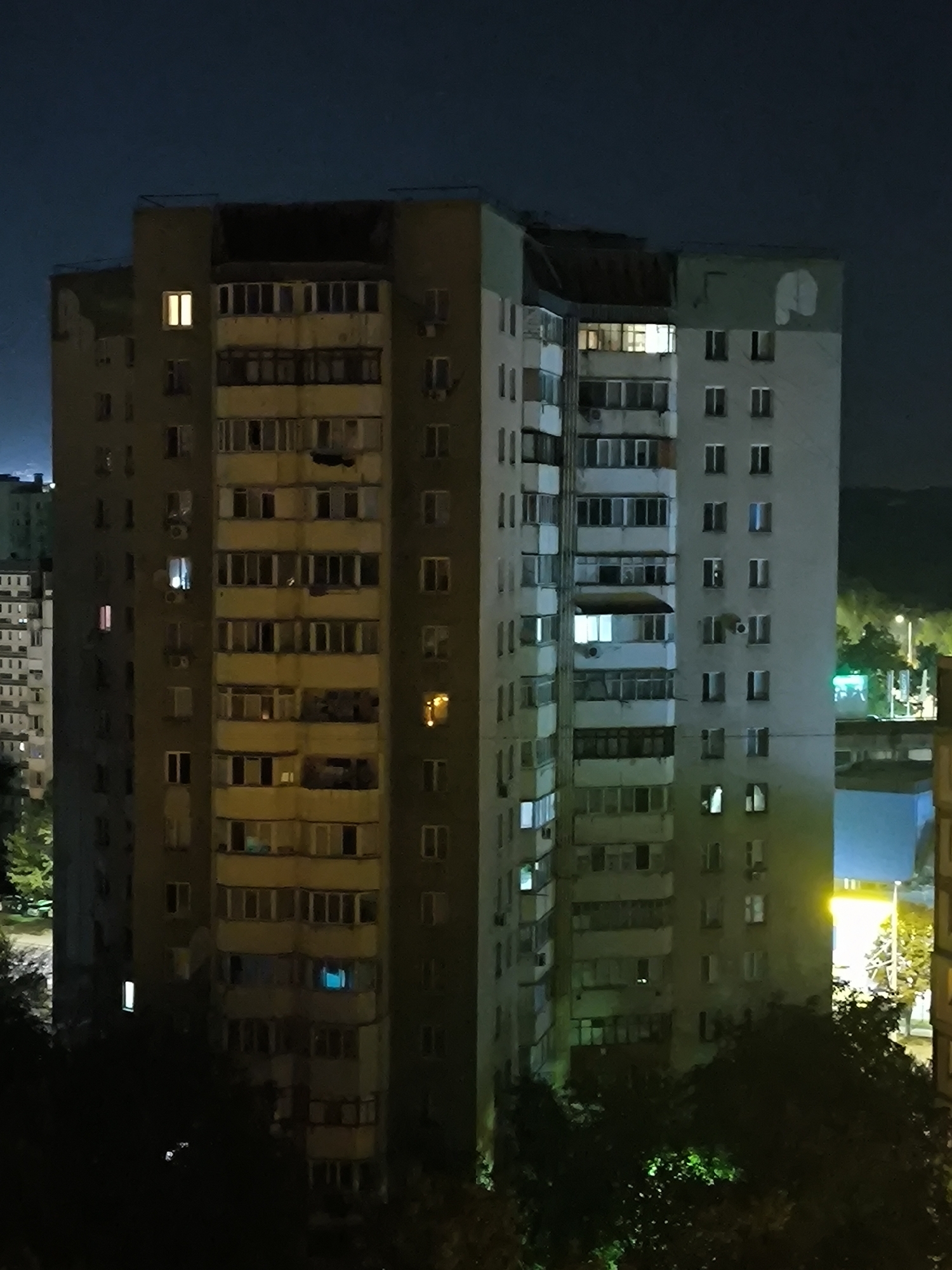 The image is a night-time, eye-level view of a tall, multi-story residential building in Chișinău, Moldova. The building is primarily light gray with sections painted in a darker gray or brown hue. It features numerous windows, many of which are illuminated with yellow or white light, indicating occupancy. Balconies are visible on multiple floors, some with railings and others enclosed with glass.
The building is situated in a densely populated urban environment. To the left of the main building, a portion of another, similar residential structure is visible, also with illuminated windows. In the background, trees and other buildings are discernible, with some lights from street lamps or other structures adding to the illumination. The sky is dark, suggesting late evening or night. The overall impression is of a typical urban apartment building at night, common in many Eastern European cities. The architecture suggests it is likely a Soviet-era residential block.