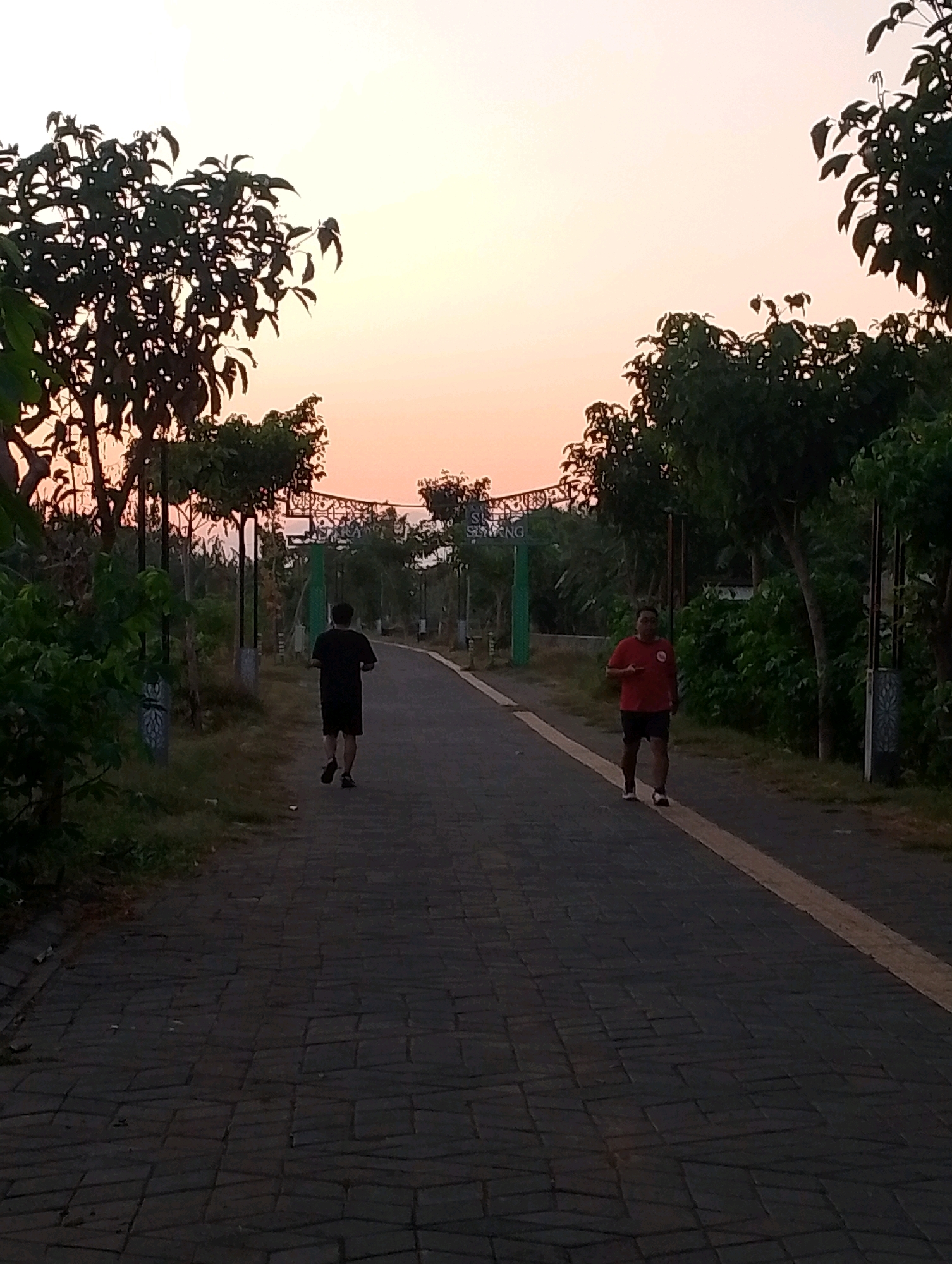 Two men are jogging on a paved walkway in what appears to be a park or recreational area at dusk. The sky is a soft orange and yellow, indicating sunset or sunrise. Trees and foliage line the sides of the walkway, with some lampposts visible. In the background, an archway with lettering, possibly signifying an entrance or landmark, is present. The man on the left is wearing a black t-shirt and shorts. The man on the right is wearing a red t-shirt and dark shorts. The walkway is made of dark grey interlocking paving stones. The overall scene suggests a peaceful outdoor activity. This location is likely the Alun-Alun Probolinggo, a central public space in Probolinggo, Indonesia, commonly used for recreation and community events, especially during dawn and dusk.