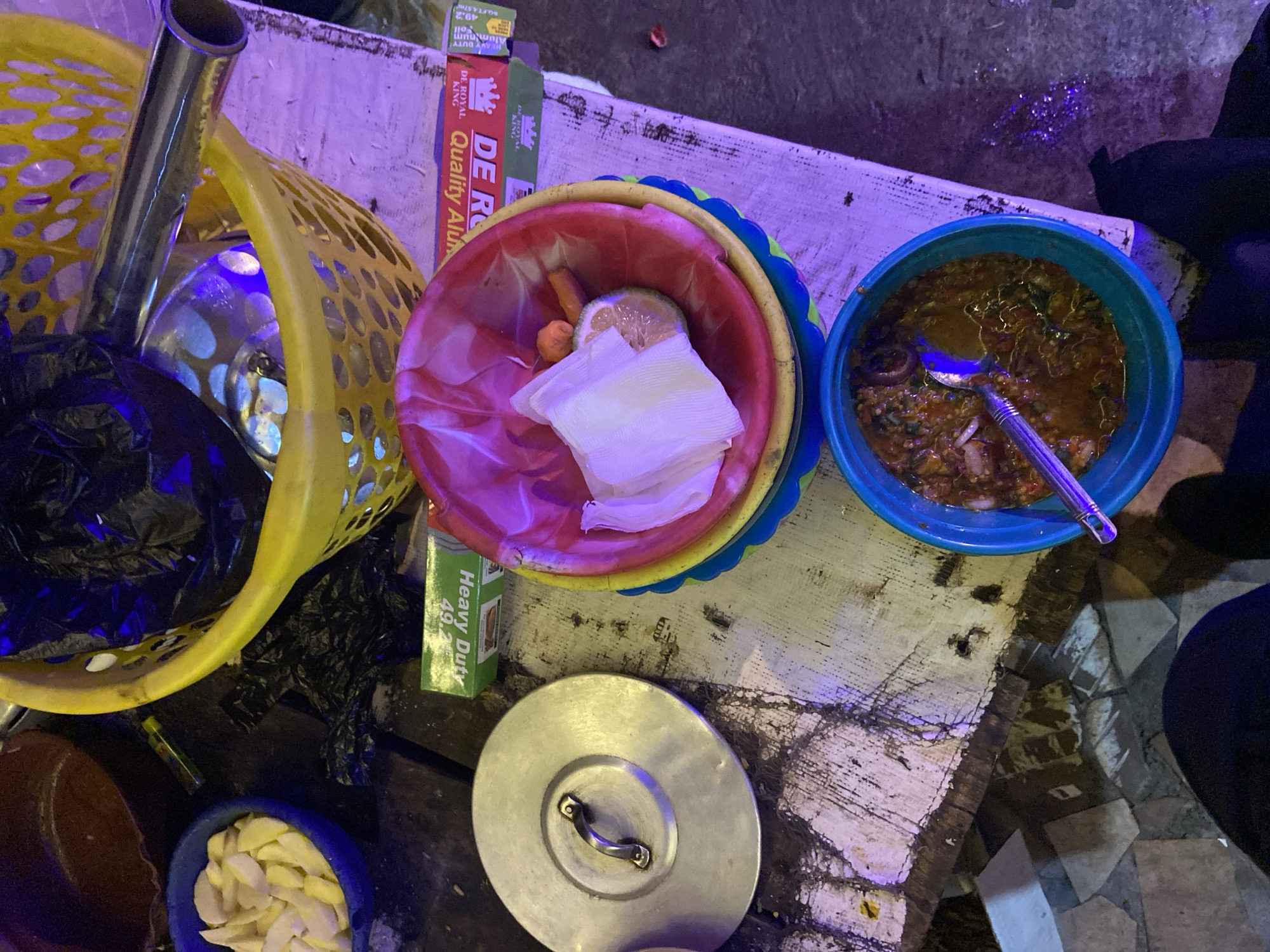 This is a photograph taken at night in Du, Nigeria, likely at a street food stall or outdoor eatery. The scene is dimly lit with a strong purple hue from artificial lighting.
In the foreground, a blue plastic bowl contains a savory, dark orange stew, possibly Egusi soup, with visible chunks of ingredients and a silver spoon resting inside. To the left, a large, circular, metallic pot lid with a handle sits on a weathered, white-washed wooden table.
Behind the pot lid, to the right, a red and yellow plastic bowl holds crumpled white napkins, a slice of lemon, and what appears to be a small orange pepper. This bowl is stacked inside a larger yellow plastic basket that contains a black plastic bag. A shiny metal pipe or rod extends from the yellow basket upwards and to the right, suggesting it might be part of a cooking setup or a stand.
A green cardboard box, likely containing aluminum foil, is visible beneath the yellow basket, with text indicating "Heavy Duty" and "Quality Alum". The table surface is rough, stained, and appears to be made of wood. Scattered debris and what look like small, white pieces of food or paper are present on the table and the ground.
In the upper left corner, a dark brown bowl or pot is partially visible. The ground appears to be a mix of concrete tiles and loose dirt, also reflecting the purple light. The image captures a snapshot of a busy, informal food preparation and serving environment.