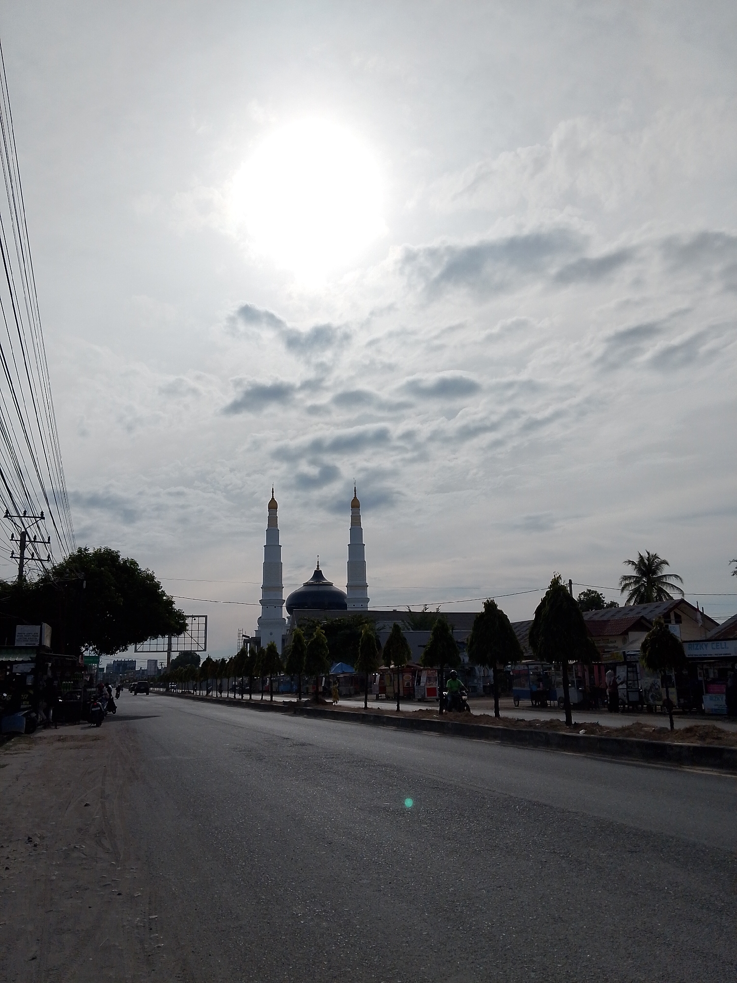 The image depicts a street scene in Pantegajah, Indonesia, with a prominent mosque in the background. The sky is overcast with bright sunlight filtering through, creating a hazy effect. A road runs through the center of the frame, lined with trees and buildings on either side. There are several vehicles on the road, including motorcycles and a car. A few people are visible, walking along the roadside and near the buildings. The mosque features two tall minarets and a dark dome. The overall atmosphere suggests a typical day in a populated urban area.