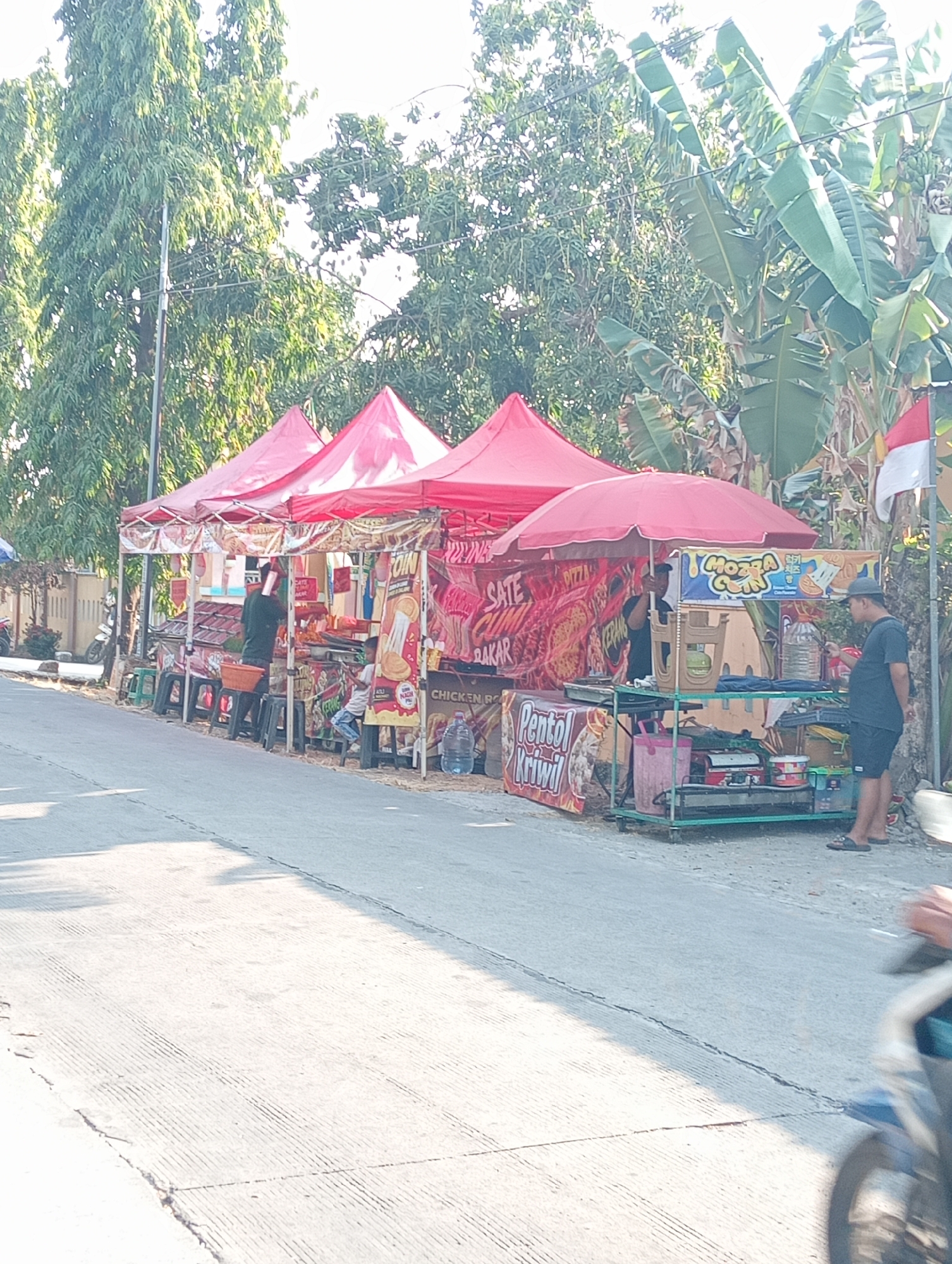 The image depicts a street scene in Tanjunganyar, Indonesia, featuring a row of food stalls under red canopies. On the left, a person in a dark shirt is standing at a stall, seemingly preparing food. To the right of this stall, a young person is seated on a stool. Further to the right, another person wearing a hat and dark shirt is standing at a stall, possibly interacting with the vendor. The stalls display various food items and signage in Indonesian. Tropical trees, including banana trees, and a street lamp are visible in the background. A motorcycle is partially visible in the foreground on the right, and a motorcycle is parked on the left side of the street. The overall environment suggests a warm, sunny day in a roadside commercial area.