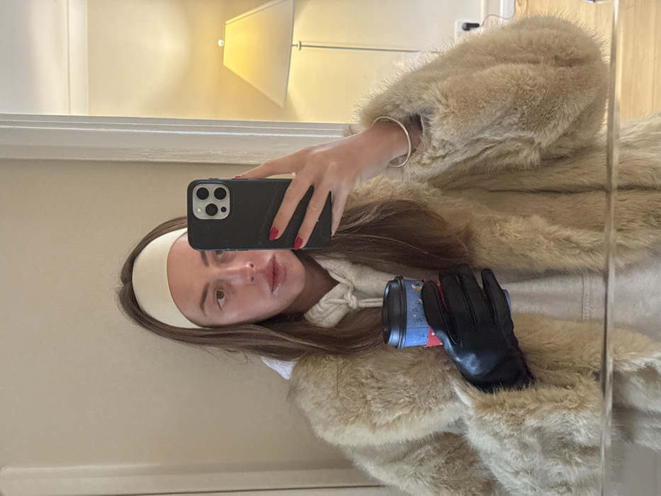 A female subject is depicted in a mirror selfie, observed from the chest up, indoors, located in the city of London, United Kingdom. She has long brown hair and wears a cream-colored headband. Her right hand, featuring red-painted fingernails and a silver bangle on the wrist, holds a black smartphone vertically to capture the image, partially obscuring the right side of her face. She is dressed in a light grey hooded sweatshirt beneath a tan faux fur coat. Her left hand, covered by a black leather glove, holds a blue and black disposable coffee cup with a white lid and a decorative graphic sleeve. The background reflection reveals a plain light-colored wall, a white door frame on the right, and an illuminated modern floor lamp with a light-colored conical shade and a metallic stand.