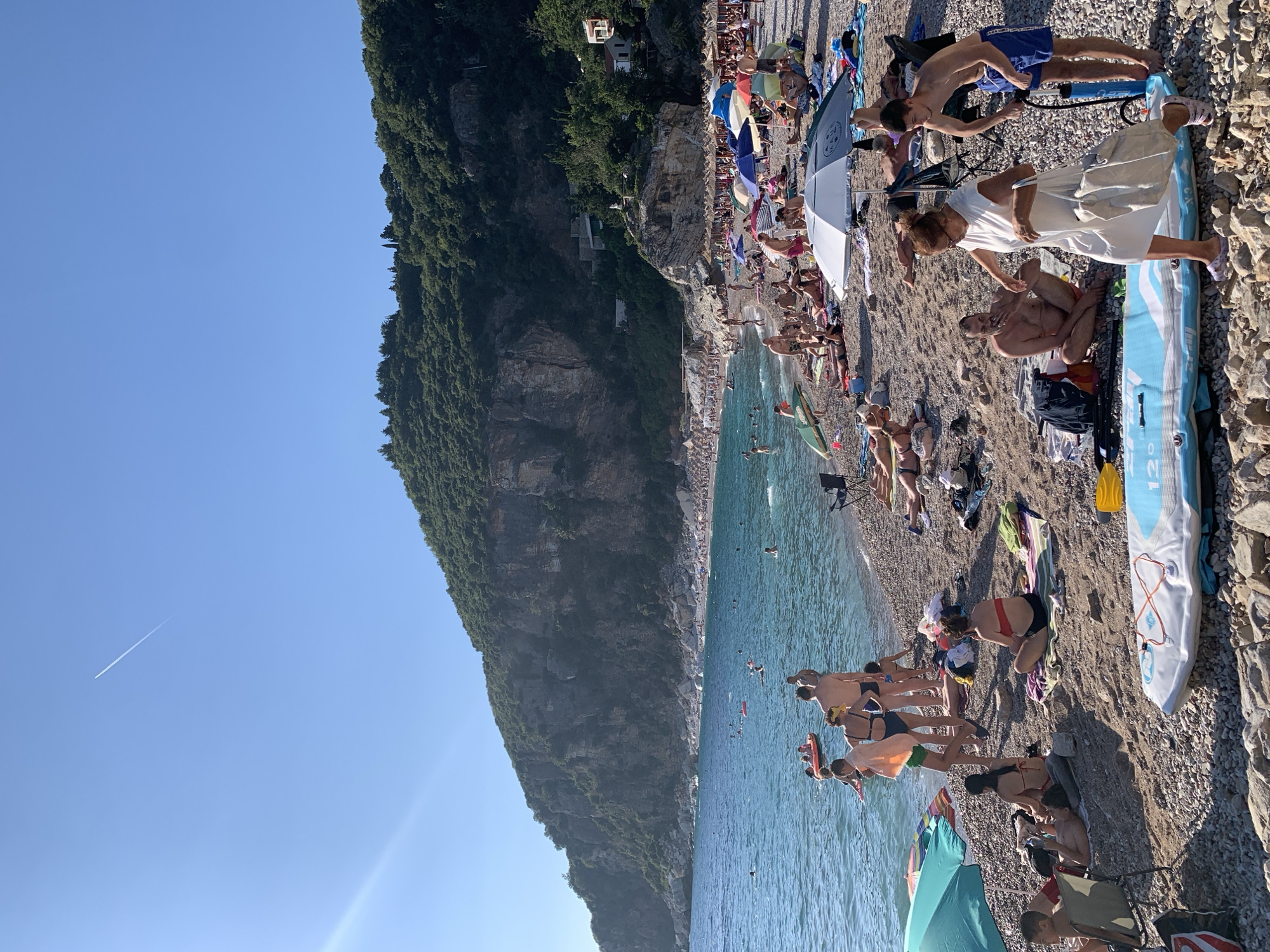 The image depicts a crowded beach in Mirište, Montenegro, under a clear blue sky. The beach is covered in pebbles, and the turquoise sea stretches out towards a backdrop of a lush, green, rocky mountain. Numerous people are visible on the beach, some sunbathing on towels and under beach umbrellas, while others are in the water, swimming or playing on inflatables. Several beach umbrellas are set up, providing shade. In the foreground, a large paddleboard is laid out on the pebbles. People are engaged in various activities, including relaxing, swimming, and interacting with each other. The overall scene captures a vibrant summer day at a popular coastal resort.