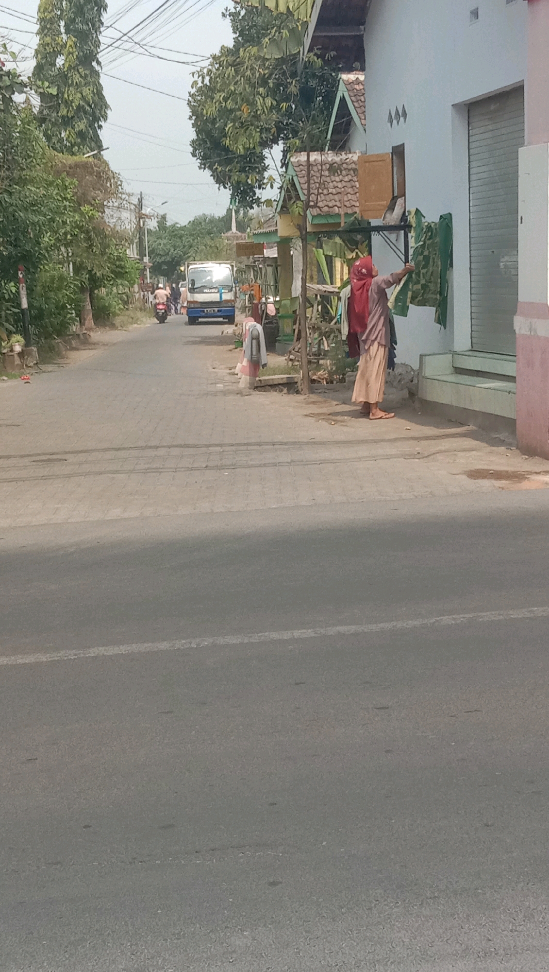 A street scene in Karang Anyar, Indonesia, features a paved road with a slight curve. On the right side of the road, a person wearing a red headscarf and a beige skirt is hanging clothes on a clothesline attached to a white building. The building has a metal roller door and a window with wooden shutters. A tree with dense green foliage is to the left of the building. On the left side of the road, there is a line of trees, some with tall, slender trunks. A blue truck with "ISUZU" branding is driving away from the camera down the road. Several other vehicles, including a motorcycle, are also visible on the road. The sky is overcast, and the overall lighting suggests it is daytime. A sidewalk made of paving stones runs along the right side of the road, with a concrete curb separating it from the road. Small plants and debris are scattered along the sidewalk.