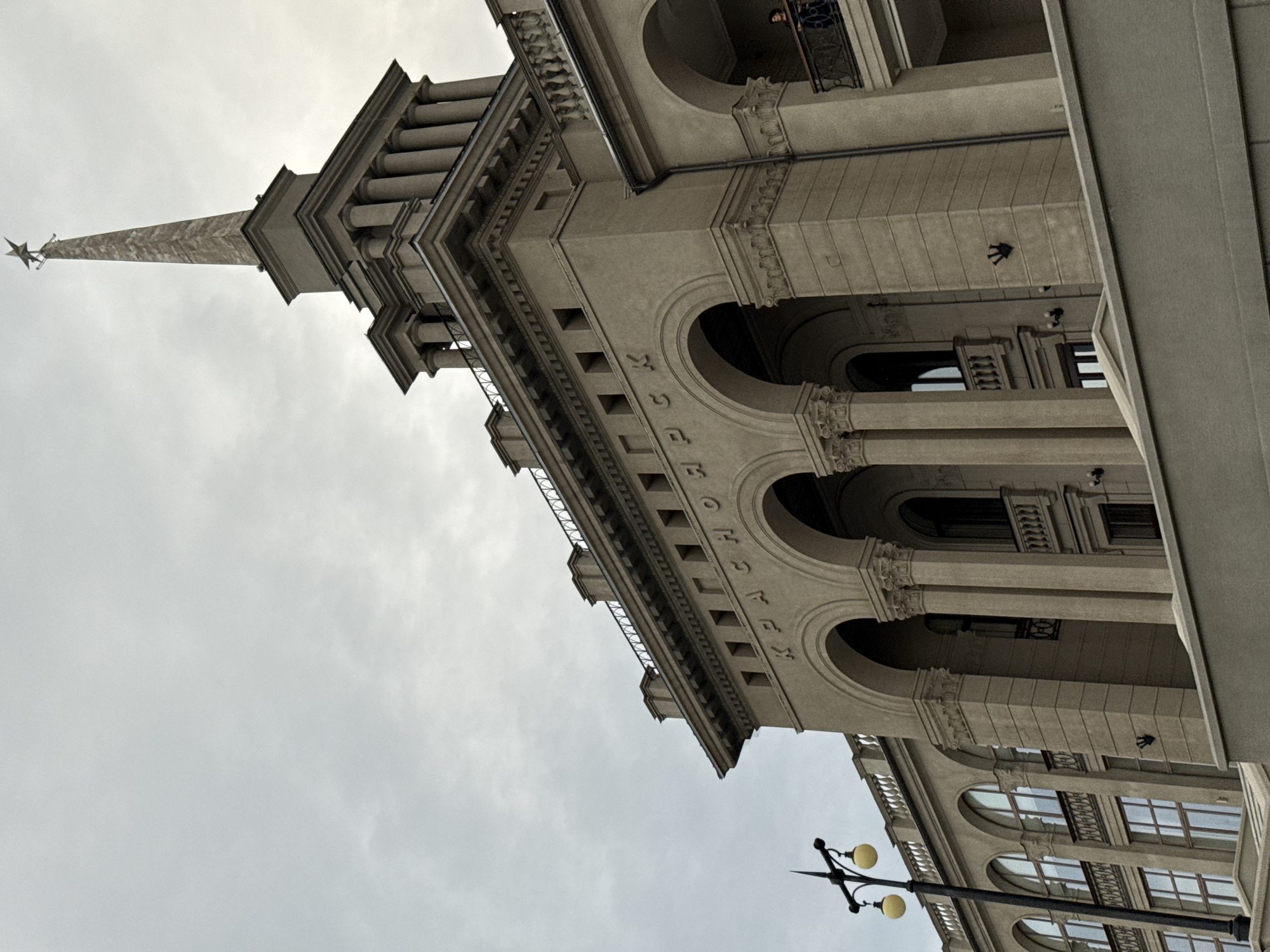The image shows the Krasnoyarsk Railway Station building in Krasnoyarsk, Russia. The building is made of stone and has a neoclassical architectural style. It features a large central portico with arches and columns, topped by a spire with a star. The building is adorned with inscriptions and decorative elements. In front of the building, there is a street lamp with two globes. The sky is overcast. A person is visible on a balcony on the right side of the building. The building is an important landmark and transportation hub in the city.
