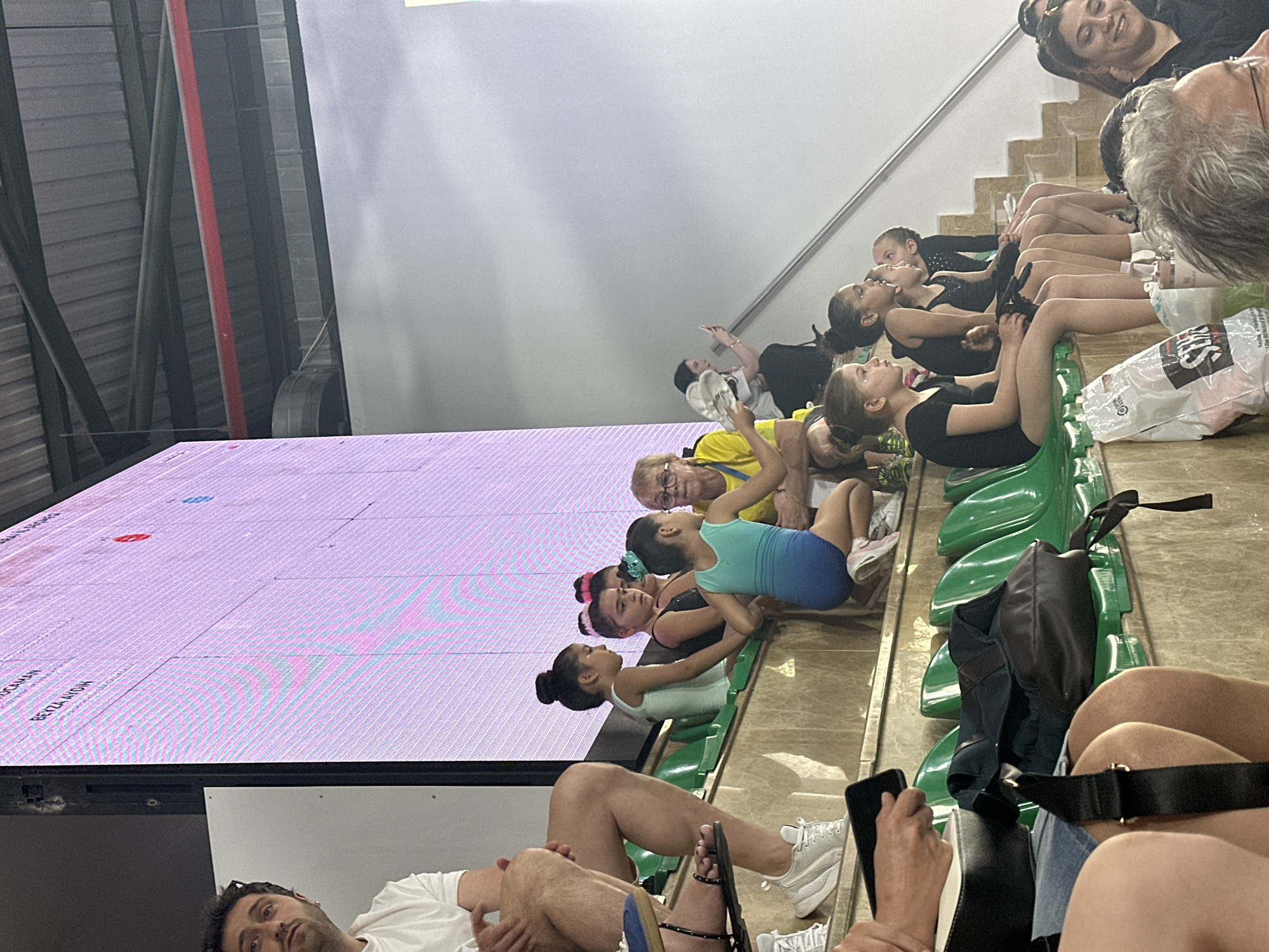 This image captures a scene from an event at the Sancaktepe, Turkey sports hall, likely a gymnastics competition, based on the attire of some participants. In the foreground, a man with dark hair and beard sits on a bench, looking up and to his left. To his right, several young girls in leotards are seated on green bleachers, some looking towards a large, illuminated screen displaying text and graphics, suggesting it's a scoreboard or information display. One girl in a turquoise leotard is closer to the viewer, interacting with an adult woman wearing a yellow top. In the background, more people are visible, some using mobile phones. The environment appears to be an indoor sports facility with tiered seating and overhead lighting structures. The focus is on the spectators and young athletes, with the large screen dominating the upper portion of the image.