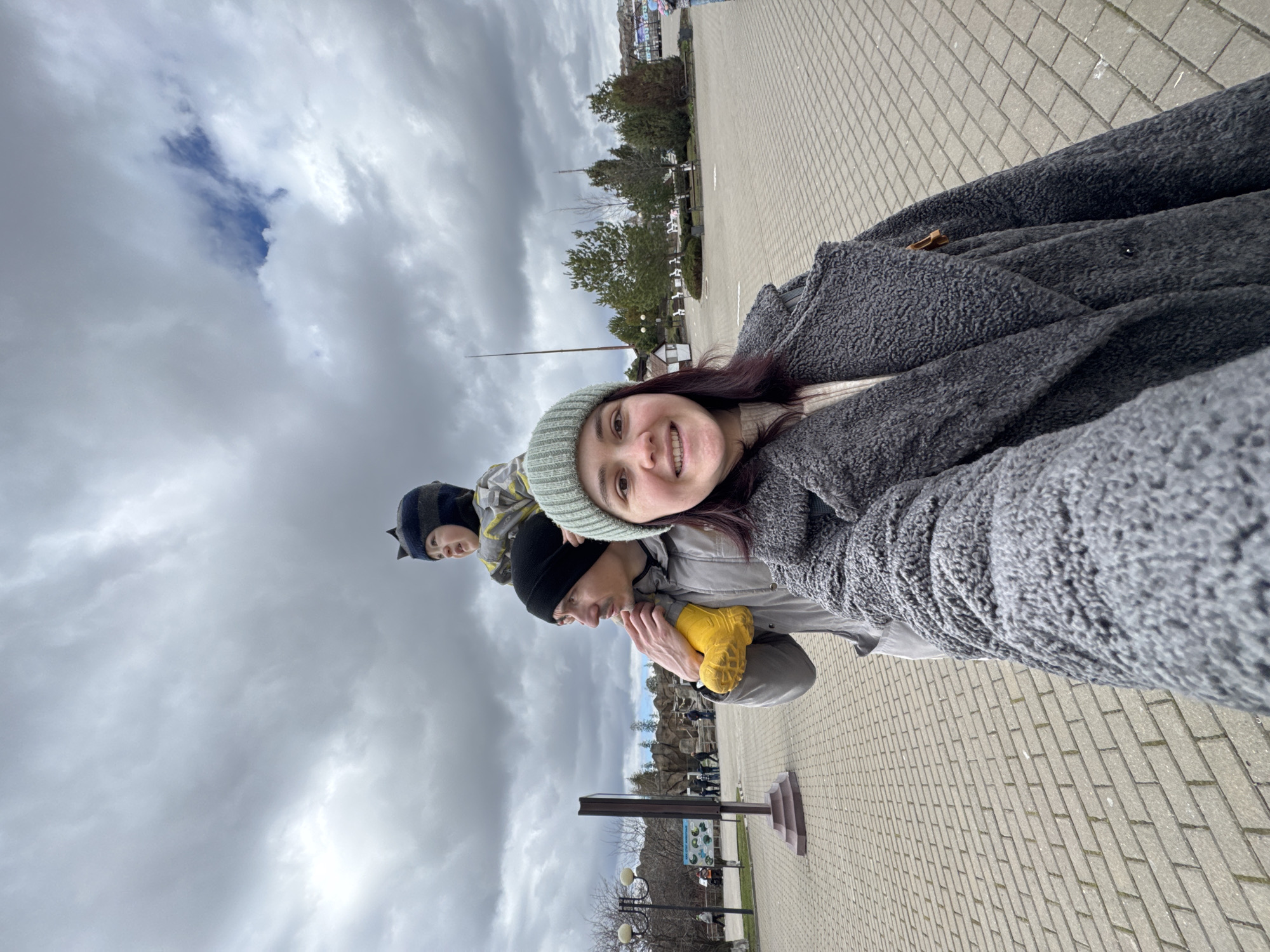 The photo is taken at the Izhevsk Zoo in Izhevsk, Russia. The image shows a woman wearing a beanie and a grey jacket taking a selfie. A man wearing a black beanie is carrying a child on his shoulders; the child is wearing a blue and grey hat with ears. The background includes paving stones, trees, a sign, and cloudy skies. Several people are walking about behind the subject.