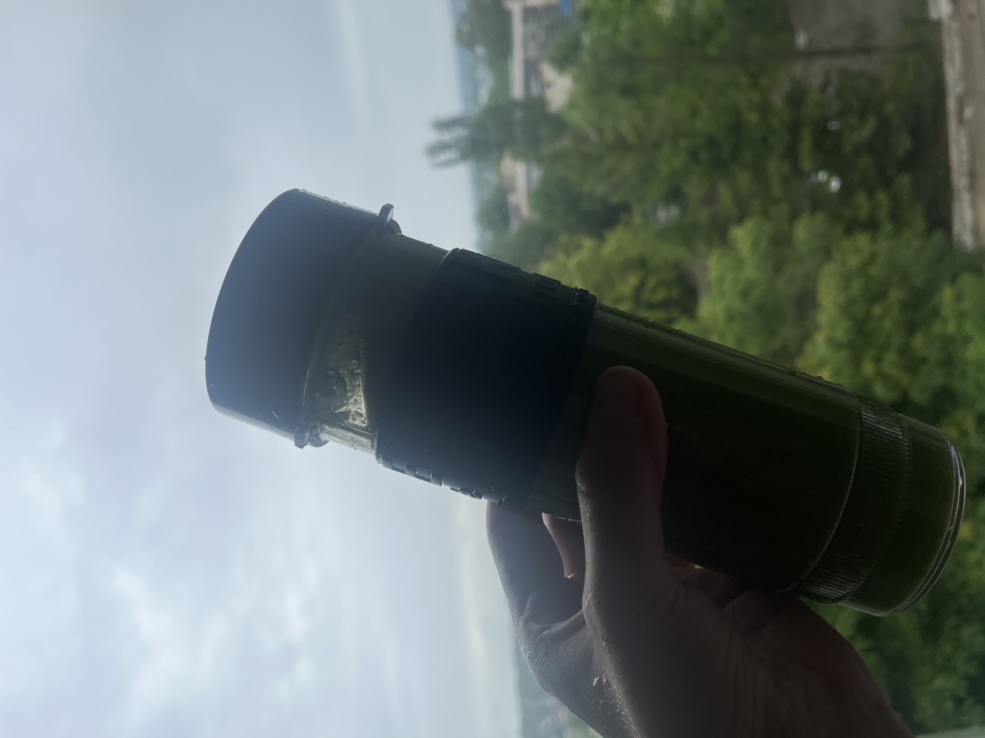 An out-of-focus, close-up shot shows a person's hand holding a dark green, clear plastic bottle filled with a dark green liquid, possibly a smoothie or juice. The bottle has a black rubber grip and a dark lid. The background is a blurry landscape of green trees and buildings under a cloudy sky, suggesting an urban environment. The lighting is diffused, indicating it might be overcast or late in the day.