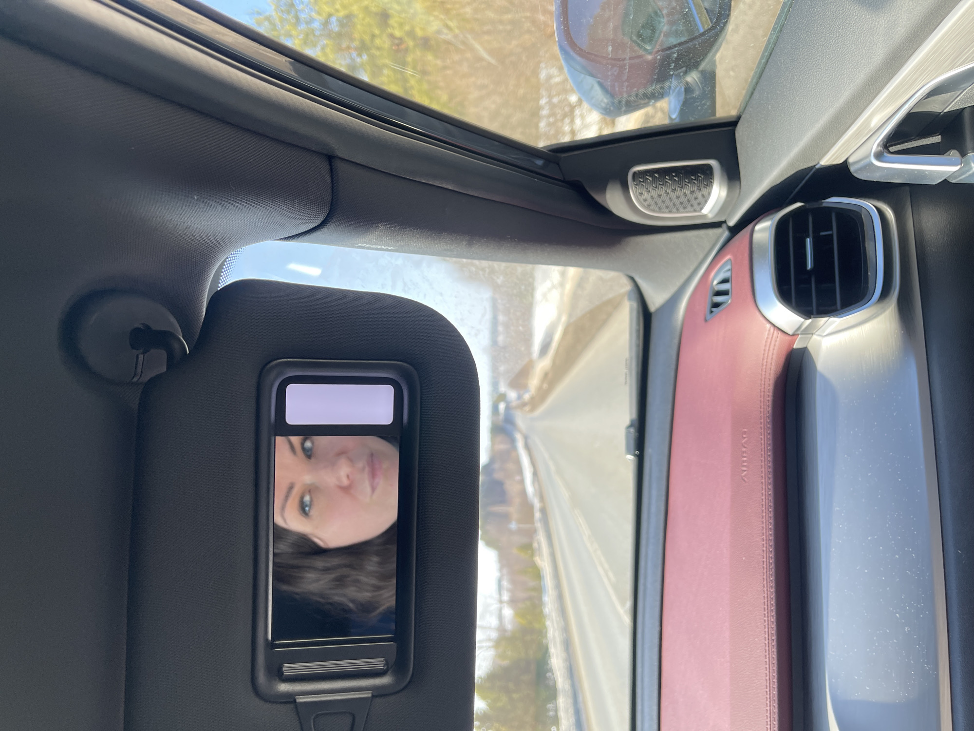 The image depicts the interior of a vehicle, viewed from the front passenger seat, moving forward on an asphalt road in Urtalga, Russia. A black sun visor is down, revealing an illuminated vanity mirror. The mirror contains the reflection of a woman with dark wavy hair and light-colored eyes, looking towards the viewer's left. A bright, rectangular white object partially obscures the right side of her reflection within the mirror. The car's dashboard is visible, featuring a maroon upholstered section with "AIRBAG" text and a lower silver-toned metallic trim with a horizontal air vent on the right. A speaker grille is integrated into the passenger door panel near the A-pillar. Through the windshield, a two-lane road recedes into the distance, flanked by partially snow-covered verges and a sparse treeline under a clear blue sky. The passenger-side mirror is partially visible on the right, reflecting the exterior environment.