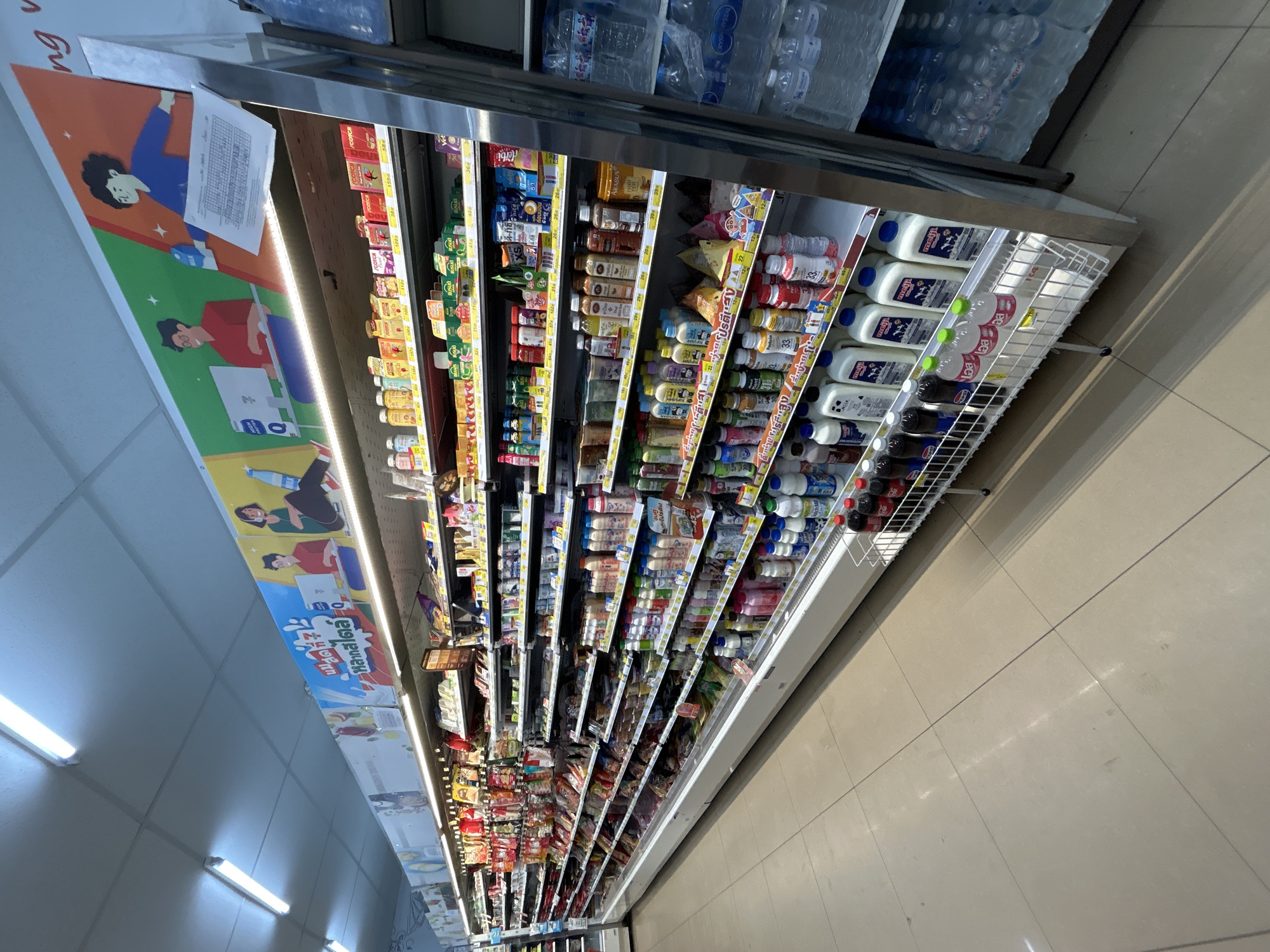 The image shows a brightly lit interior of a convenience store, likely a 7-Eleven, located in Baan Hua Thanon, Thailand. The perspective is a wide shot from an aisle, looking towards the front of the store and a refrigerated section on the right. The ceiling is white with fluorescent lighting panels. A large, colorful advertisement banner stretches across the top of the shelves, depicting animated figures and text in Thai.
The shelves are fully stocked with a variety of products. The middle and upper shelves display packaged goods such as drinks, snacks, and other convenience items. Prominently featured are numerous bottles of milk and other beverages on lower shelves and in a wire basket at the end of an aisle. These bottles are in various sizes and brands, with many exhibiting colorful labels. A refrigerated display case on the right side of the image contains stacked bottles of water. The floor is tiled in a neutral, light beige color. No people are visible in the scene. The overall impression is of a well-organized and well-stocked retail environment.
