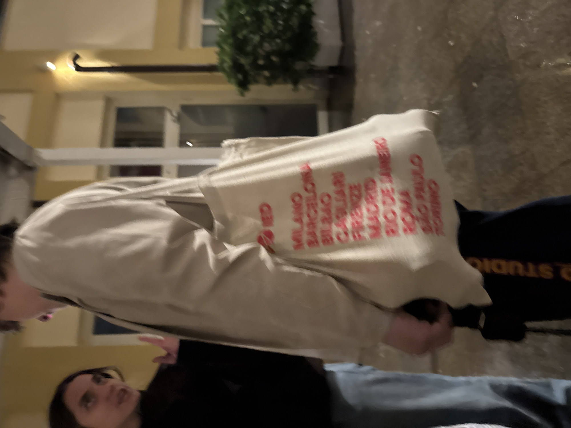 The image shows a person wearing a beige jacket and carrying a tote bag printed with "IED" and a list of cities including "MILANO" and other locations. Another person in a black shirt and jeans is partially visible in the lower portion of the frame. The background shows a building with a light fixture, windows, and a potted bush. The city location is Milan, Italy, as indicated on the tote bag.
