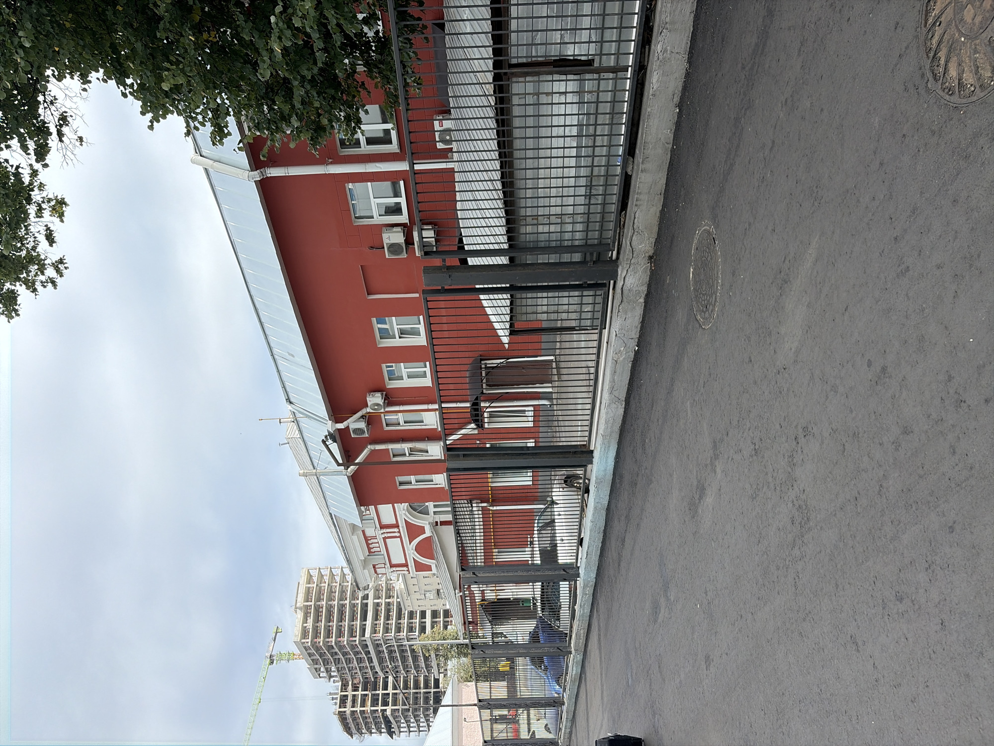 The image shows a street view in Moscow, Russia, on a cloudy day. In the foreground, there is an asphalt road with two manhole covers. A black metal fence with vertical bars runs along the right side of the road. Behind the fence, there is a red building with white-trimmed windows and air conditioning units mounted on the exterior walls. Part of the roof of the red building is visible, covered with a light-colored metal. To the left of the red building, a multi-story building under construction with a crane is visible in the background. Several cars are parked behind the fence, some of them partially obscured. Trees with green foliage are visible in the upper right corner of the image. The sky is overcast.