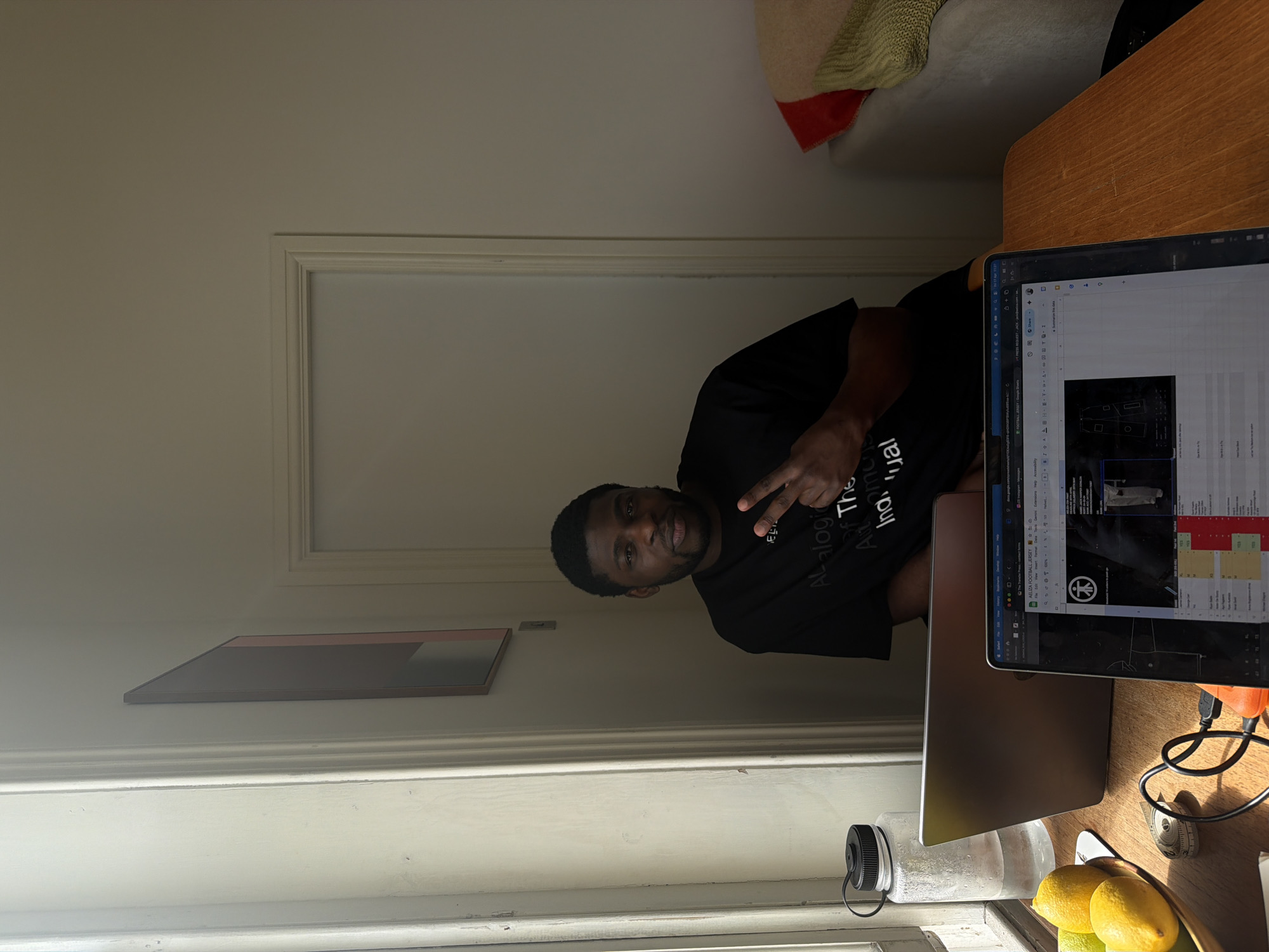 This media file captures an indoor scene, likely in London, United Kingdom, showing a man seated at a wooden desk with a laptop. The man, who has dark skin, short dark hair, and a beard, is looking directly at the camera with a slight smile and making a peace sign gesture with his right hand. He is wearing a black t-shirt, which has visible text including "EL", "Analogic", "Of The", "Aut", "Ind?mone", and "ual" (potentially forming "Analogic Of The Individual").
On the desk in front of him is an open silver laptop, displaying a screen with various elements including a spreadsheet and what appears to be design or product imagery related to "AELIZA FOOTBALL JERSEY". Other text visible on the screen includes "FOOTBALL JERSEY - Google", "PRESS REQUEST JACK", and "Share". To the man's left, a clear plastic water bottle is present, and to his right, a bowl containing lemons and limes.
The room has light-colored walls, with white molding or trim visible, possibly around a doorway or window on the left side of the frame. A framed artwork featuring abstract geometric shapes in muted tones (grey, pinkish-orange) hangs on the wall behind him. In the background, to the right, a portion of a sofa or chair with a light green and red throw or blanket can be seen. Strong natural light, possibly from a window to the left, illuminates the scene, suggesting it is daytime. The man appears friendly and engaged, pausing his work to pose for the photograph.