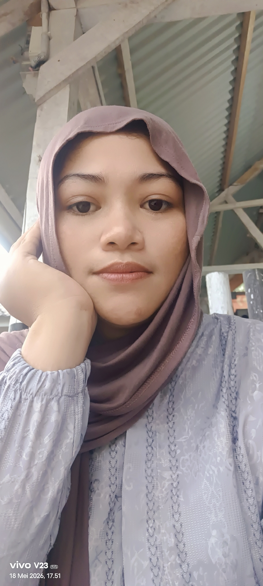 A woman wearing a brown hijab and a light blue patterned blouse is shown in a close-up selfie. She is resting her face on her left hand. The background includes elements of a building structure, with light-colored supports and corrugated metal. The lower left corner of the image includes a timestamp: "vivo V23 5G 18 Mei 2026, 17.51". The location cannot be determined as Lhokseumawe from the image alone.