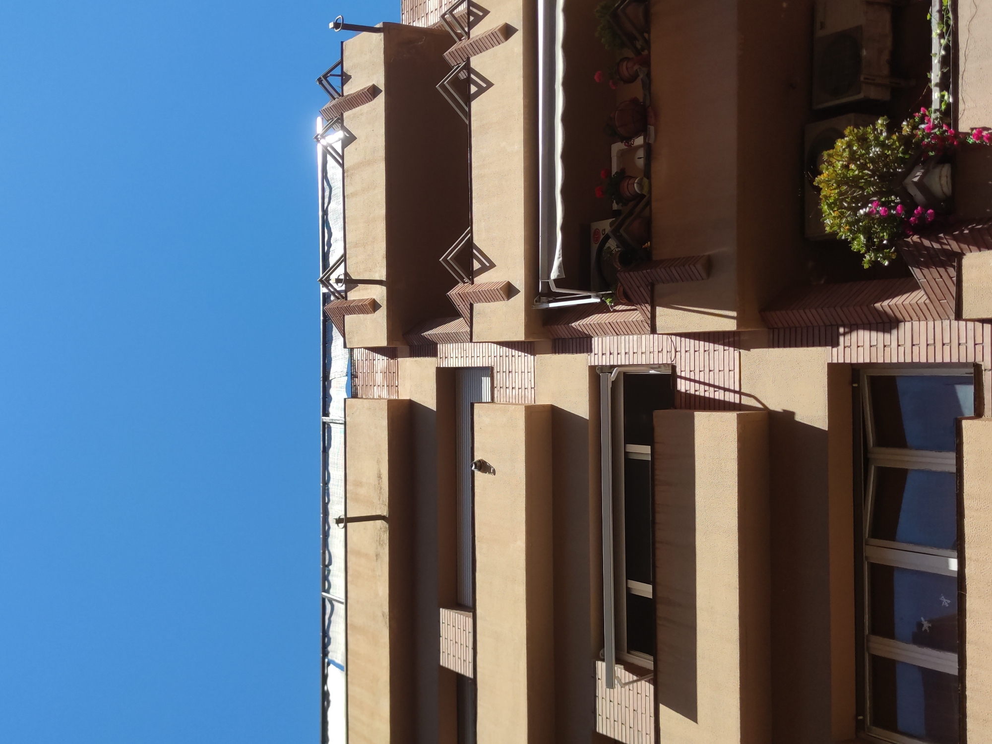 Viewed from a low angle, the tan-colored side of a building stands against a bright blue sky. The building's architecture features vertically stacked rectangular protrusions and balconies. Some balconies have plants. Windows are visible, with one open. Brick accents and metal railings add detail. The angle suggests the building is tall, with sunlight highlighting the structure. The location is not discernible from the image.