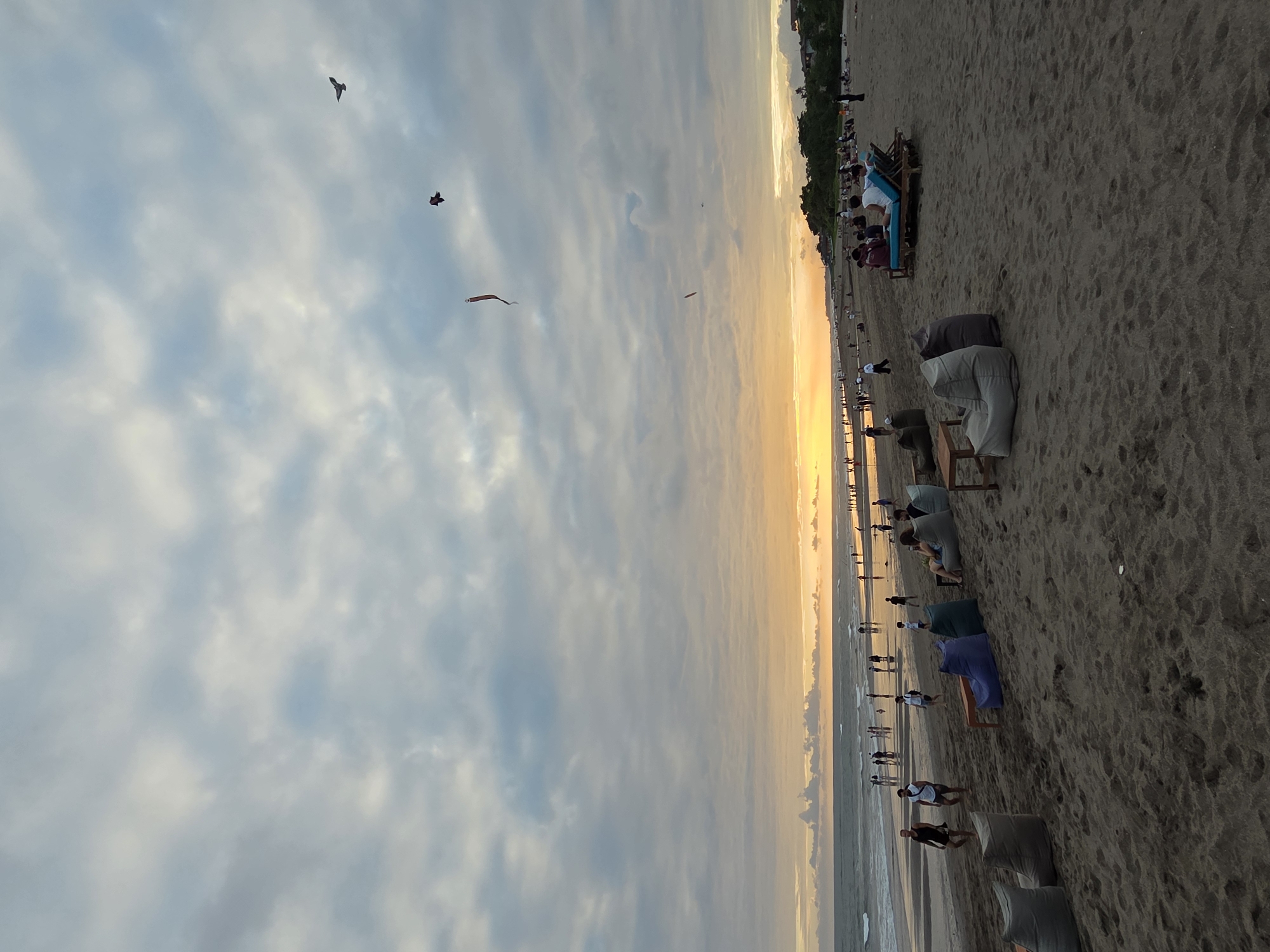 This is a wide-angle, full shot of a beach scene in North Kuta, Indonesia, during sunset. The sky is filled with a mix of grey and white clouds, with the setting sun casting a warm orange glow on the horizon. The ocean is calm, with small waves rolling towards the shore.
On the beach, numerous people are visible. Some are walking along the waterline, while others are gathered in groups, seated on beanbag chairs or lounging on beach chairs. There are several beanbag chairs scattered across the foreground and mid-ground of the image, in shades of grey, purple, and brown. Two lounge chairs with a blue cushion are occupied by people. A few small tables are placed near the seating areas.
In the sky, two kites are flying, one red and one dark brown. A bird is also visible flying high in the sky. The beach is made of dark sand, and there are some sparse areas of vegetation visible in the distance, along with buildings. The overall atmosphere appears relaxed and leisurely, with people enjoying the evening by the sea.