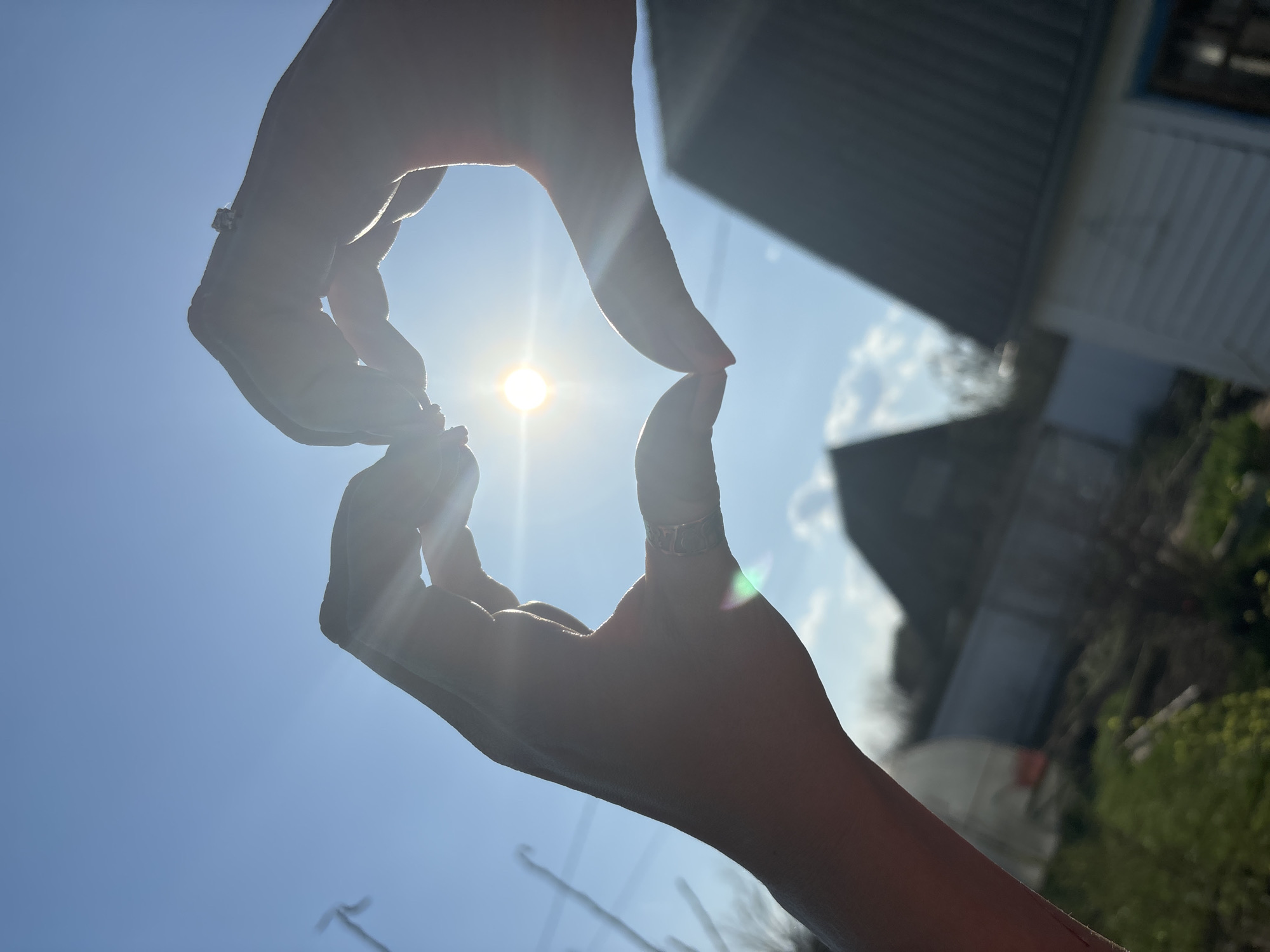 The image shows two hands forming a diamond shape with the sun at the center. The hands have rings on their fingers. In the background, there is a house and some trees against a bright blue sky with some clouds. The location appears to be outdoors during the day. The city of Noviy, Russia is not evident in the image.