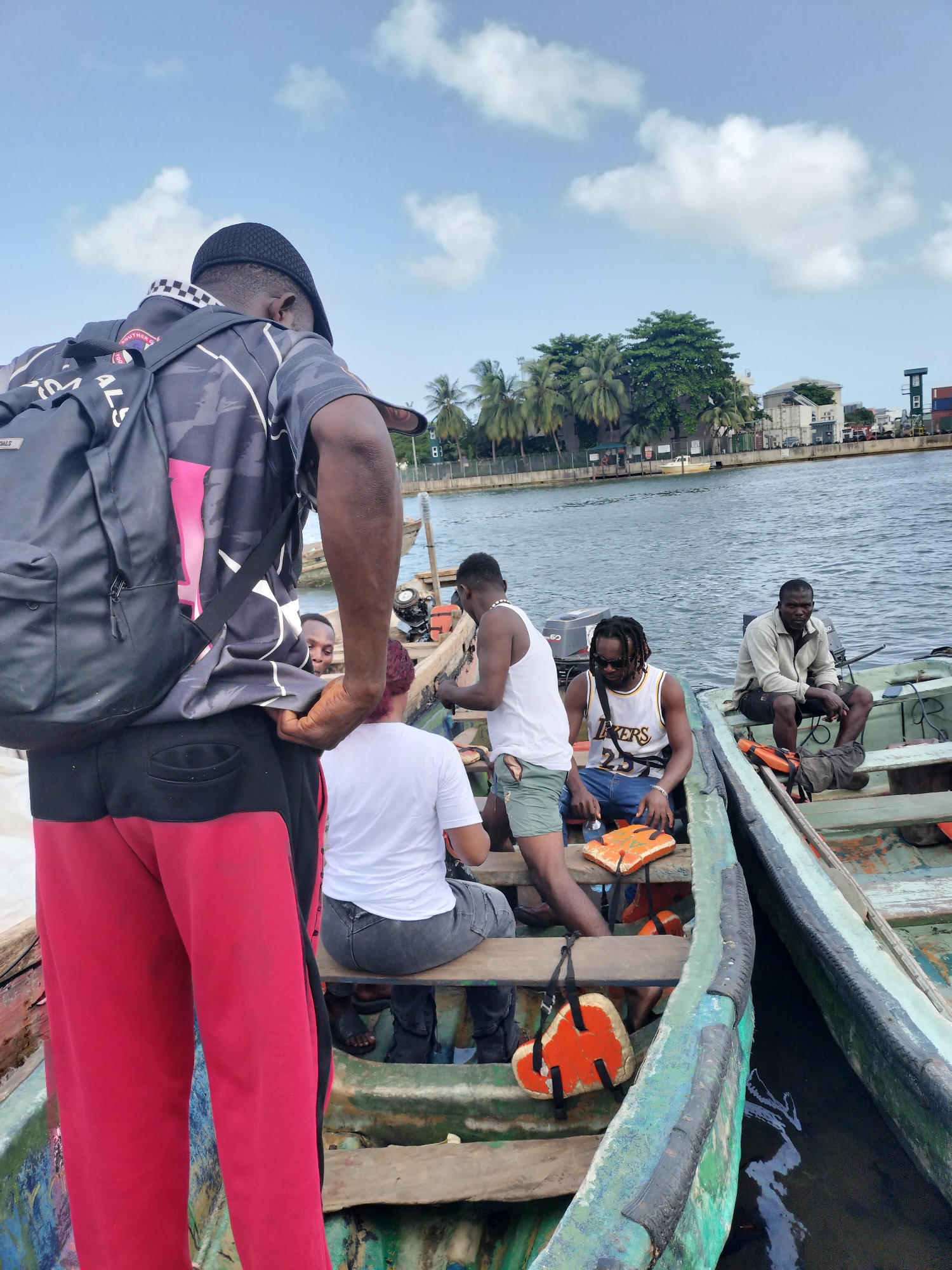 A group of six men is gathered in and around two small, open wooden motorboats on a calm waterway, likely a river or canal, under a bright blue sky with scattered white clouds. The scene suggests preparation for water travel or an ongoing journey.
In the immediate foreground, a man stands in a boat, his back to the viewer. He wears a black cap, a dark grey and pink patterned short-sleeved shirt with a backpack, and red and black sweatpants. "ESSENTIALS" is visible on his black backpack. He appears to be looking down at others in the boat.
Inside the same boat, three other men are seated. One has reddish-brown hair and wears a white t-shirt. Another, in a white tank top and green shorts, is partially visible. The third, seated centrally, wears a white "LAKERS 23" basketball jersey, blue jeans, and sunglasses, with an orange buoyant cushion on his lap. Several other orange life jackets or cushions are visible in the boat.
To the right, an adjacent, similar wooden motorboat holds another man, wearing a light-colored jacket/hoodie and dark shorts, seated and looking towards the first boat. Both boats have outboard motors. The boats show signs of wear, painted in shades of green and blue.
The background reveals the opposite bank of the waterway, lined with lush green tropical vegetation, including palm trees, and several buildings. Some structures appear industrial, possibly including shipping containers, hinting at a commercial or port area. The overall atmosphere is casual and relaxed on a bright day in what is identified as Alaiyabiagba, Nigeria.