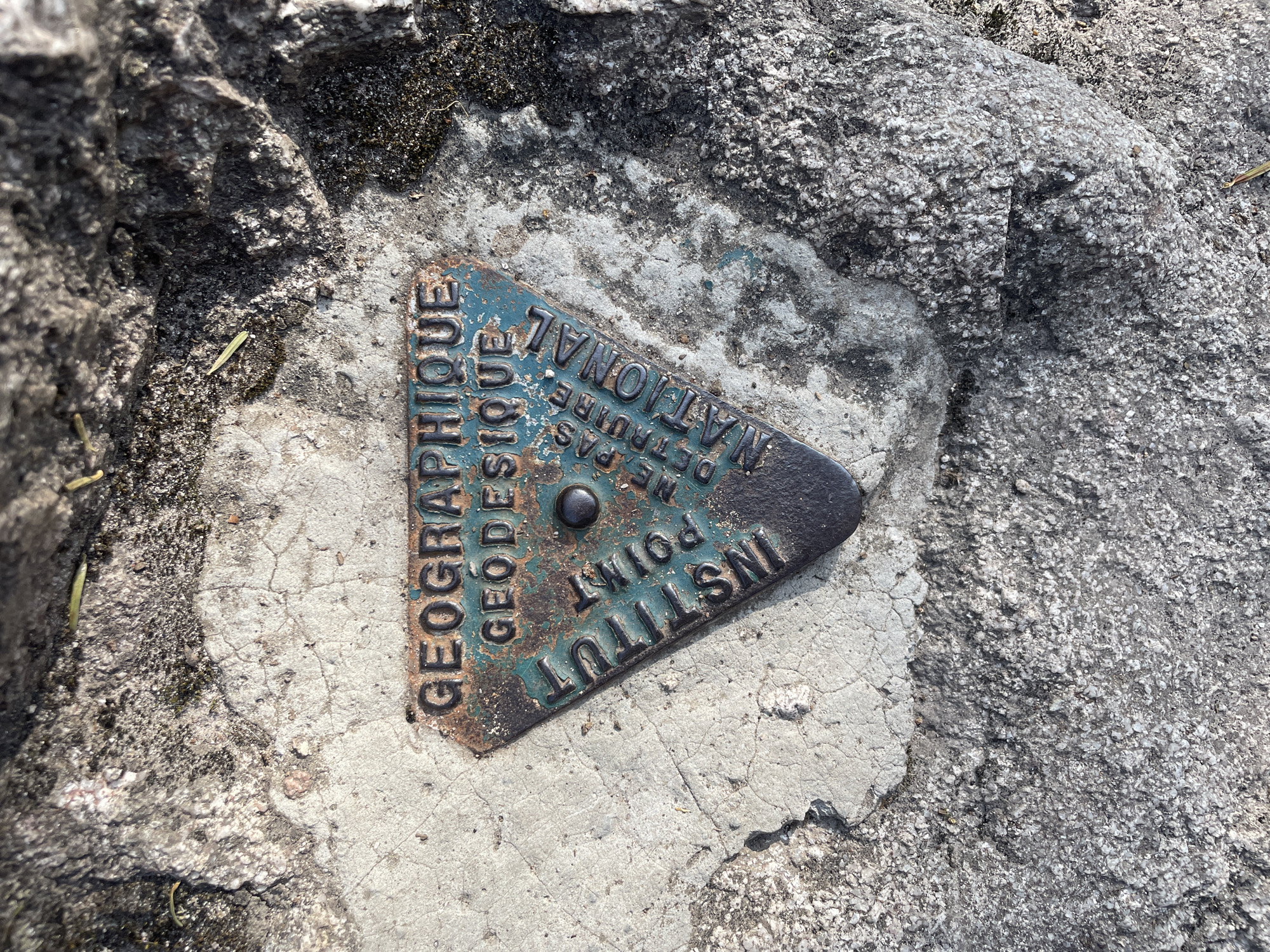 A triangular metal marker, branded with the words "GEOGRAPHIQUE GEODESIQUE INSTITUT NATIONAL" and "POINT", is embedded in a rough, grey, concrete surface. The marker is worn and partially rusted, with some of its teal-colored enamel chipped away. The surrounding environment appears to be an outdoor rocky or mountainous area, with moss and small plant debris visible on the concrete and rocks. The marker is positioned at an angle within the frame, casting a slight shadow. This marker is likely a geodetic control point, part of the French National Geographic Institute's survey network. It is located in the city of Gérardmer, France, a commune in the Vosges department.
