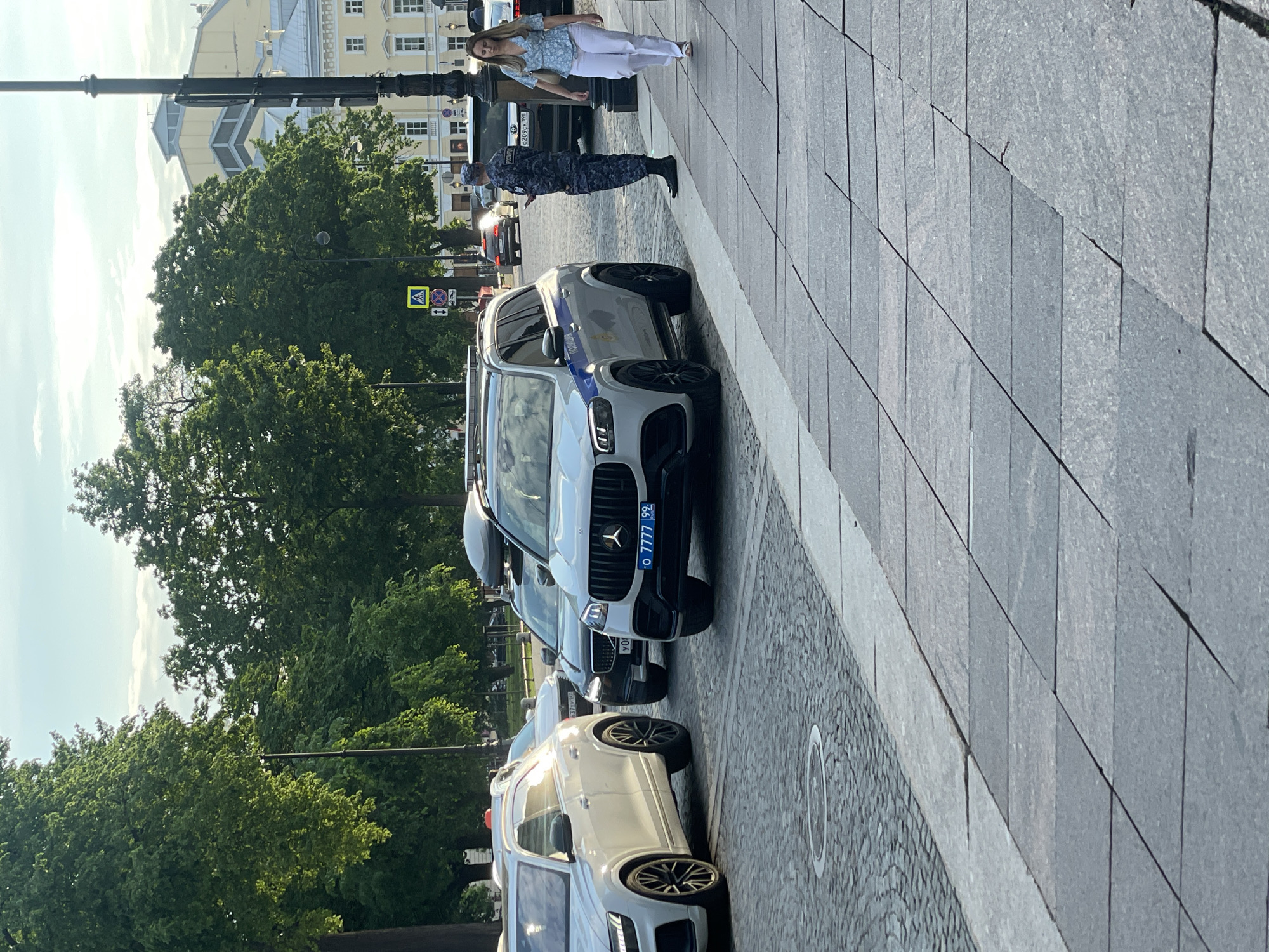 This image captures a bright daylight street scene in what is identified as Saint Petersburg, Russia. The perspective is slightly tilted, looking down a wide sidewalk and street lined with parked cars and trees.
In the foreground, a woman wearing white pants and a light blue patterned top stands on the grey stone-tiled sidewalk, looking towards the left. Beside her, a person in a dark blue/grey camouflage uniform, consistent with Russian police or security forces, stands near a white Mercedes-Benz SUV. This SUV features a blue stripe along its side, common on official vehicles, and displays a Russian license plate: "O 7777 99". The "99" code typically signifies the Moscow region, suggesting this vehicle may be from there.
Behind the Mercedes, other cars are parked in a line. One car further down has a partially visible license plate "C207 CK 198", with "198" being a Saint Petersburg regional code. Green trees with full foliage line the street, obscuring parts of the background buildings. Above the trees, a clear, bright sky is visible.
In the background, traditional-style buildings with multiple windows and pitched roofs can be seen, one notable for its yellow walls and white trim. Street furniture includes dark lamp posts and traffic signs, such as a pedestrian crossing sign and a "no stopping/parking" sign. The sidewalk is paved with large rectangular grey stone tiles, bordered by a section of grey cobblestones. No specific event is unfolding beyond people standing and cars parked in what appears to be a normal urban setting.