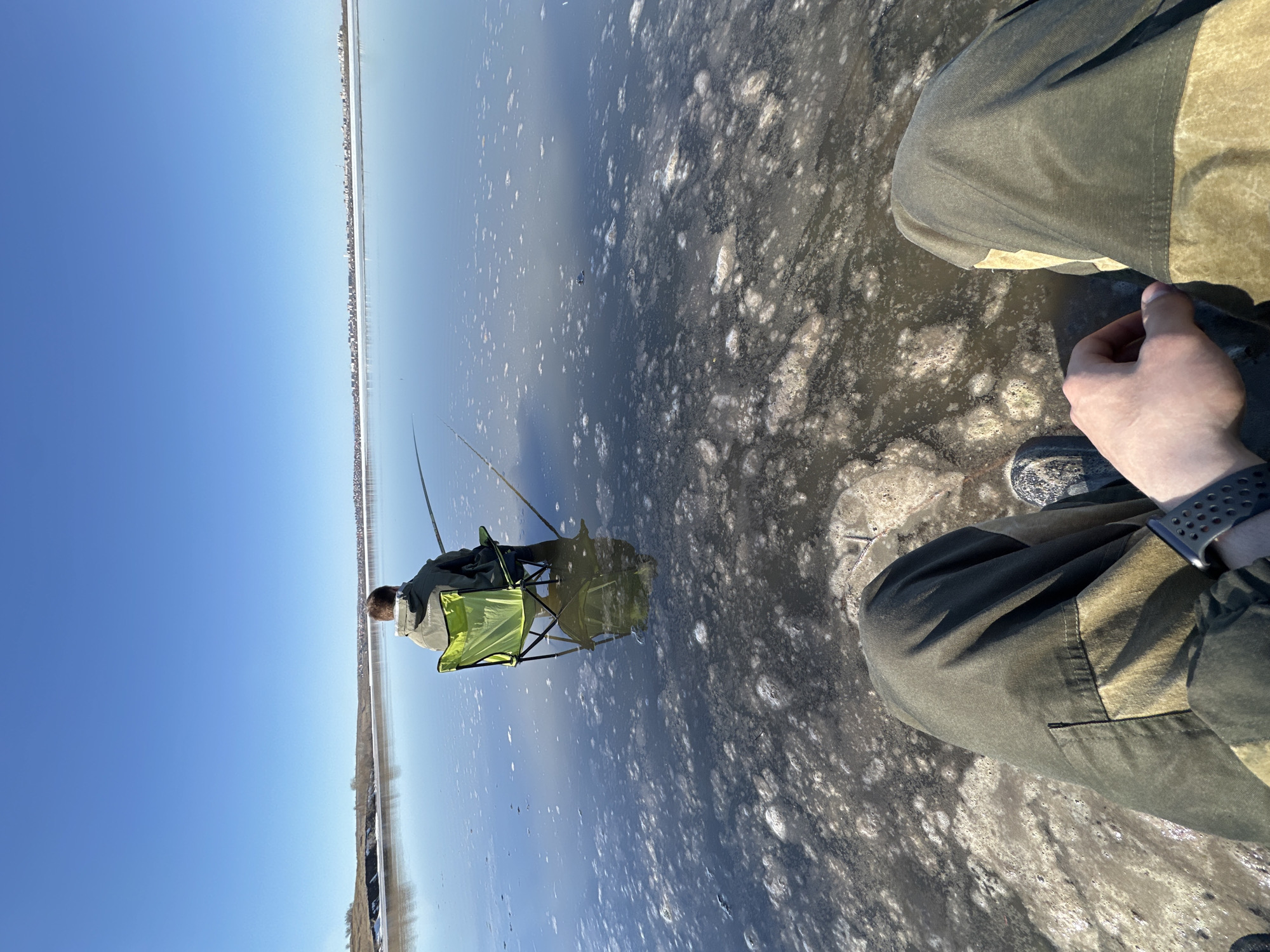 A landscape shot, taken at an unknown location in an unknown city, reveals a body of water, possibly a lake or sea, partially covered with a mix of clear and opaque surfaces. An individual is seated in a green folding chair, presumably fishing, as fishing rods are set up nearby. A second person's legs are visible in the foreground, wearing pants and a watch. The sky is clear blue, suggesting a sunny day. The terrain in the distance hints at the possibility of a skyline or land formation.