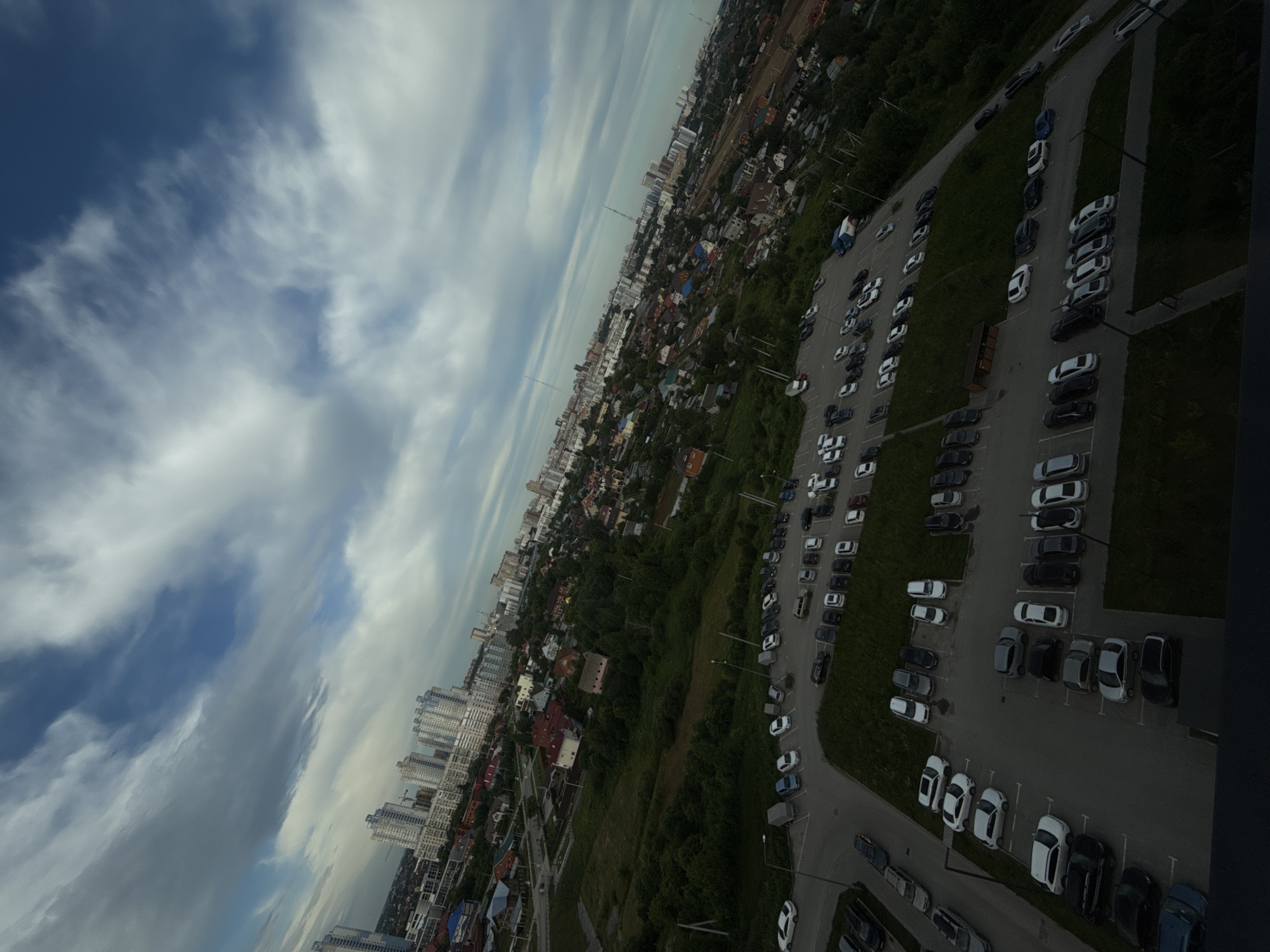 This is an aerial, wide-angle, tilted shot of a suburban area in Perm, Russia, under a cloudy sky. The scene is dominated by a mix of residential buildings and a large parking lot filled with cars. In the background, modern apartment buildings and older, smaller houses are visible, suggesting urban development. The foreground features a large, asphalt parking lot with numerous vehicles parked in designated spots. The cars vary in color and model, indicating a diverse range of ownership. There are no people clearly visible in the image. The environment appears to be a typical residential area, with green spaces and trees interspersed among the buildings and parking areas. The overall impression is a snapshot of everyday life in a Russian city.