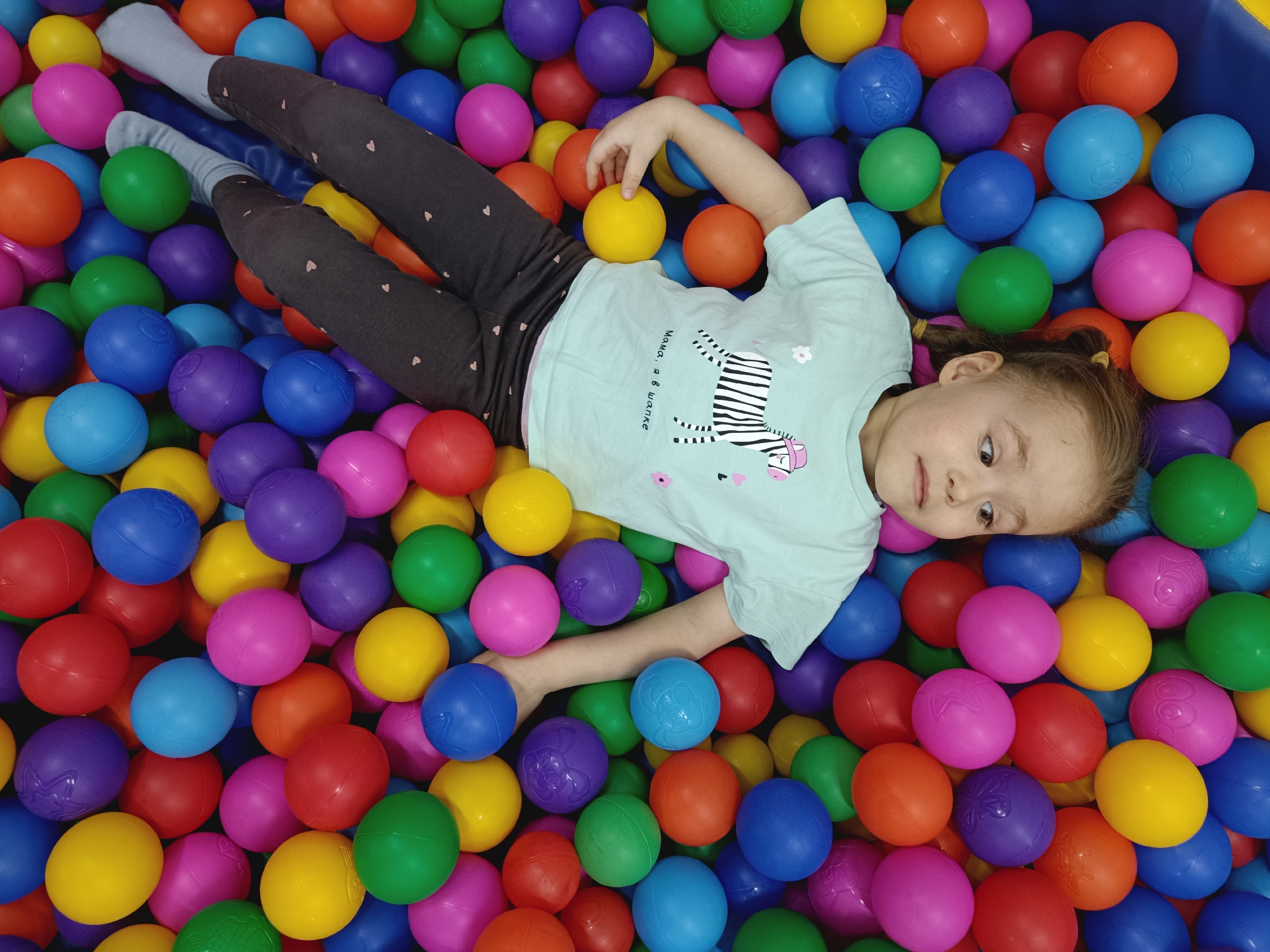 A young girl is lying in a ball pit filled with multicolored plastic balls (red, orange, yellow, green, blue, and purple). She is wearing a light blue t-shirt with a zebra design and dark gray pants with small heart patterns. The girl is holding a yellow ball in one hand and a blue ball in the other. Her hair is pulled back in a ponytail. The location is an indoor play area, presumably in the city of Unknown, Unknown.
