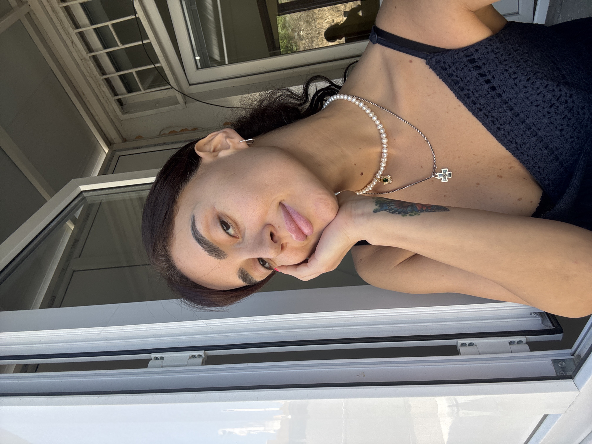The image shows a close-up of a woman resting her cheek on her hand as she looks towards the camera. She has dark hair pulled back and is wearing a dark top, a pearl necklace with a green pendant, and a silver necklace with a cross pendant. She also has a tattoo on her left arm. The woman is positioned near an open window with white framing. The background shows part of a building with a window and outdoor greenery visible through it. The scene appears to be an outdoor setting, possibly a balcony or terrace of a residential building. It is daytime and the lighting suggests it is sunny. There is no explicit indication of the city being Tbilisi, Georgia.