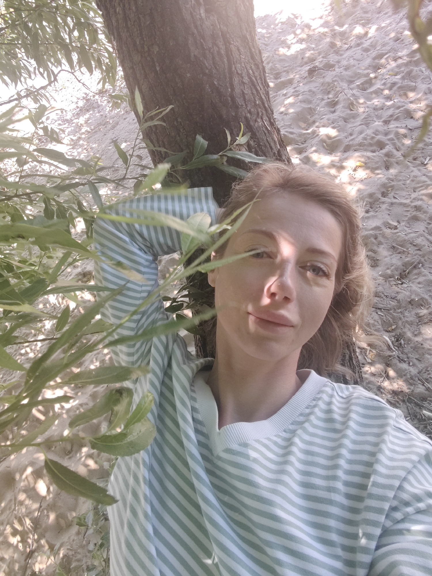 A light-skinned woman with blonde, wavy hair lies on her back, propped against the base of a tree. She wears a light-colored sweatshirt with thin, horizontal green stripes. Her left arm is bent at the elbow, with her forearm resting on her chest. Her right arm is not visible. Sunlight filters through the leaves of a tree, casting dappled shadows on her face and the sandy ground. The background consists of sand with scattered dry leaves and twigs. The woman is in the foreground, taking a selfie. The location is a sandy area with trees, possibly a park or beach, consistent with areas found within Saint Petersburg, Russia.