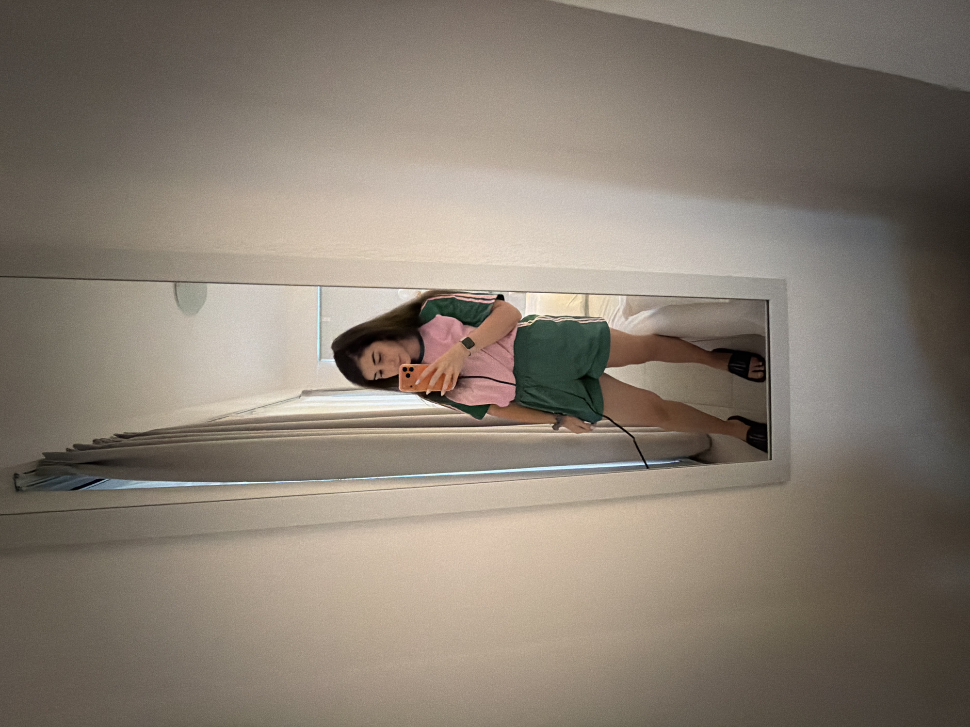 A woman is taking a mirror selfie in a room. She is wearing a pink and green athletic-style outfit with striped details on the sleeves and shorts. She has dark hair and is holding an orange smartphone. She is also wearing black slide sandals. The room appears to be a hotel room, with a neutral-colored wall, a curtain, and a bed visible in the reflection. The mirror is tall and narrow and is framed by a white border. The lighting is soft and diffused.